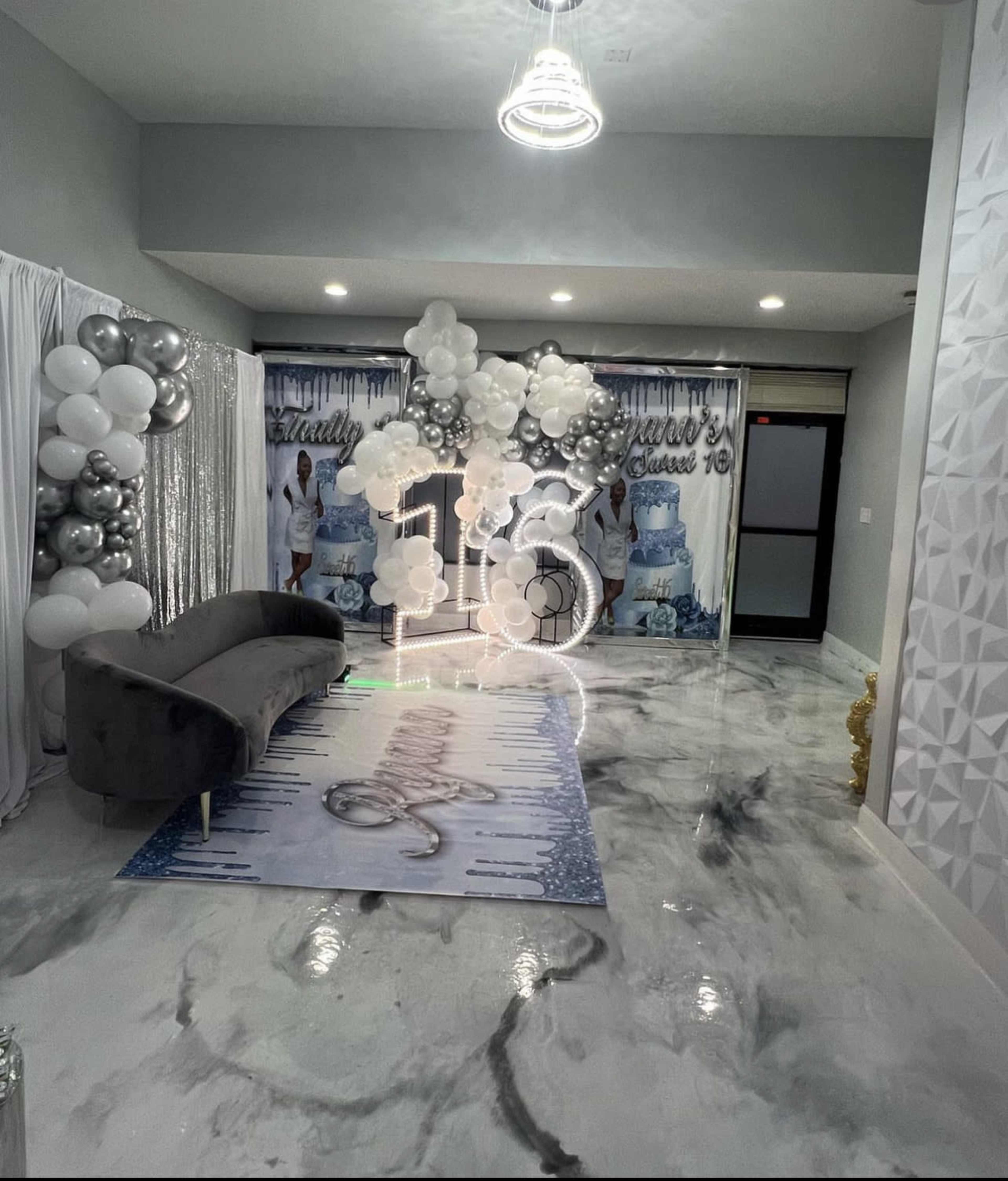 A decorated event space with a silver and white color scheme, featuring a large "16" display and a backdrop with a blue cake theme.