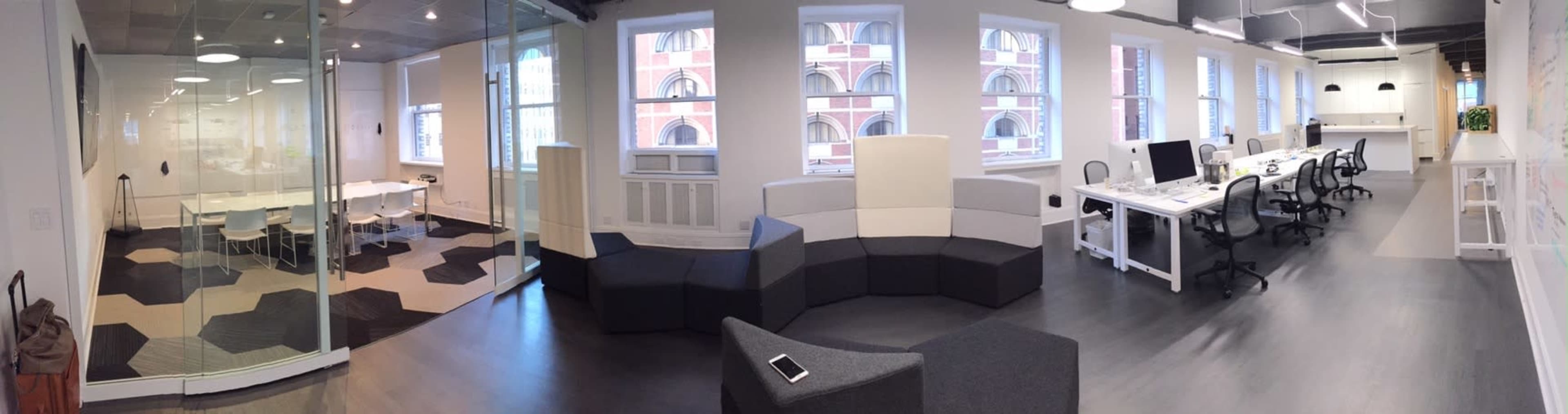 A modern office space featuring a glass-walled meeting room, a collaborative work area with desks and chairs, and a seating area with modular couches.