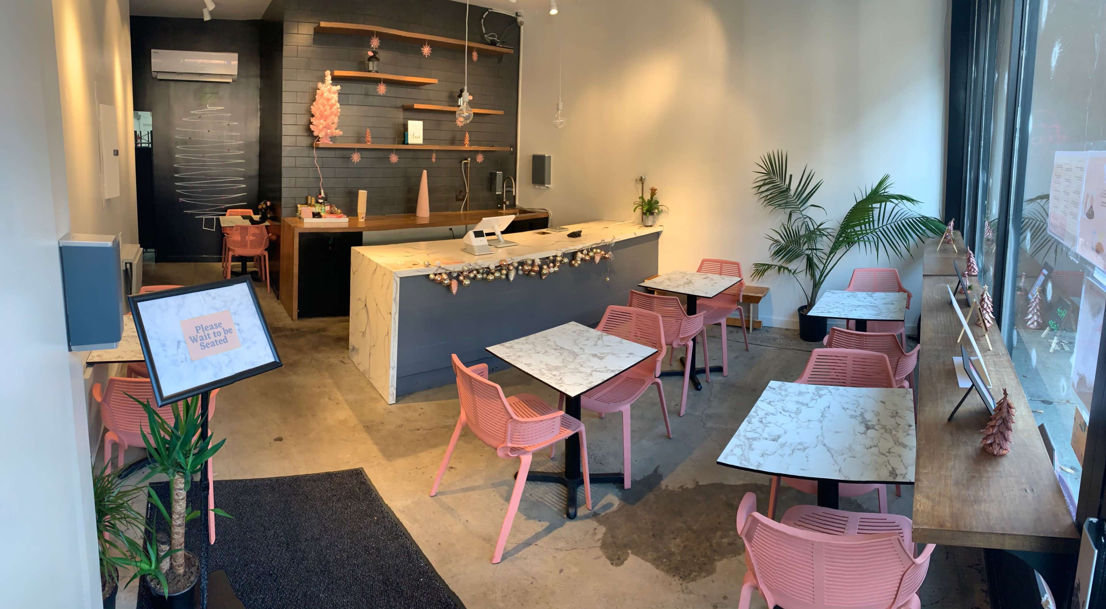 A modern cafe interior featuring marble-topped tables with pink chairs, a counter with decor, and plants in the corners.