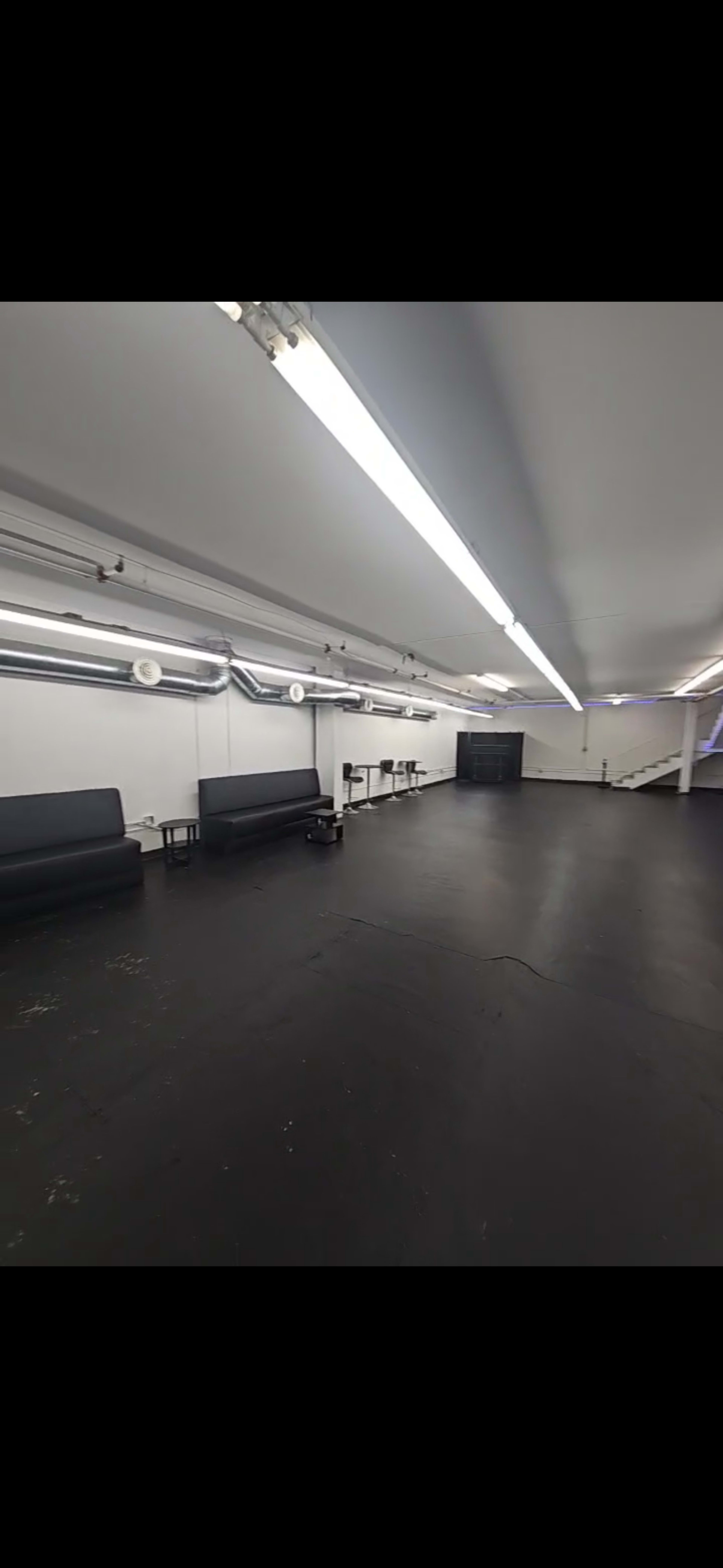 The image shows a spacious, empty room with black flooring, fluorescent lighting, and a few benches along the walls.