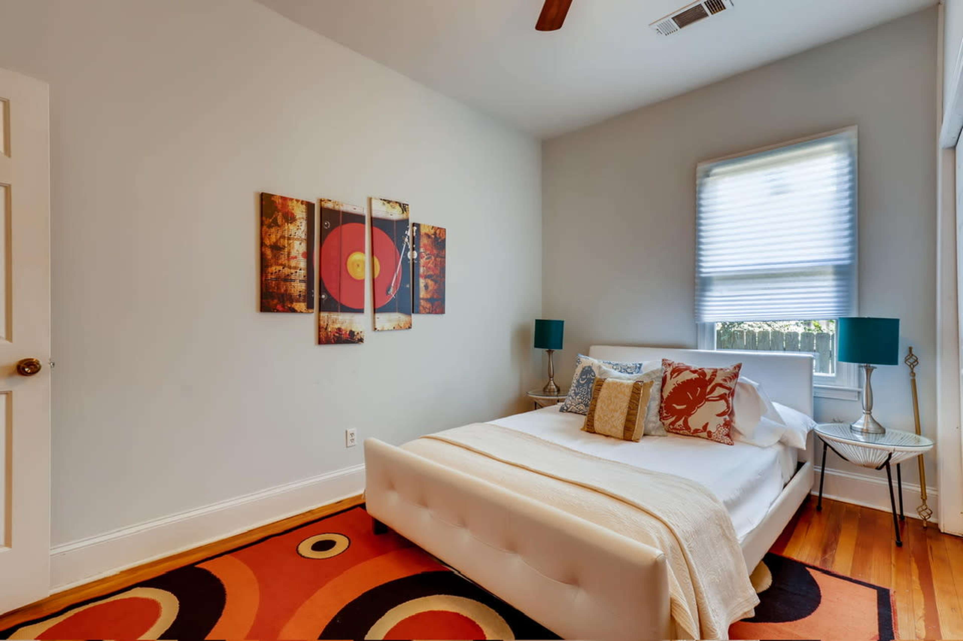The image shows a compact bedroom with a white bed, decorative pillows, and colorful artwork on the walls, accompanied by a patterned rug and blue lamps on nightstands.