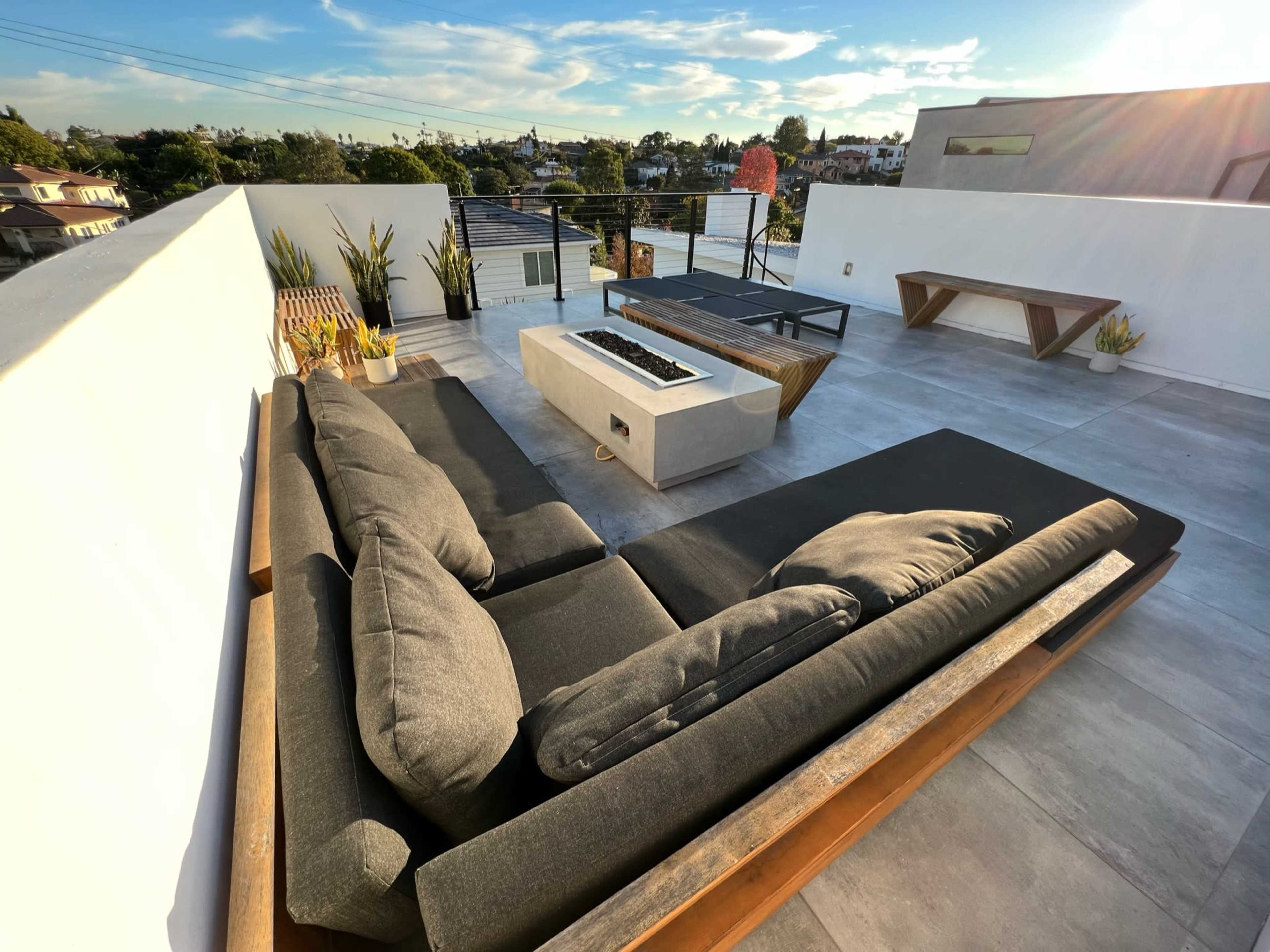 The image shows a modern rooftop terrace featuring a sectional sofa, a fire pit, and wooden furniture surrounded by potted plants.