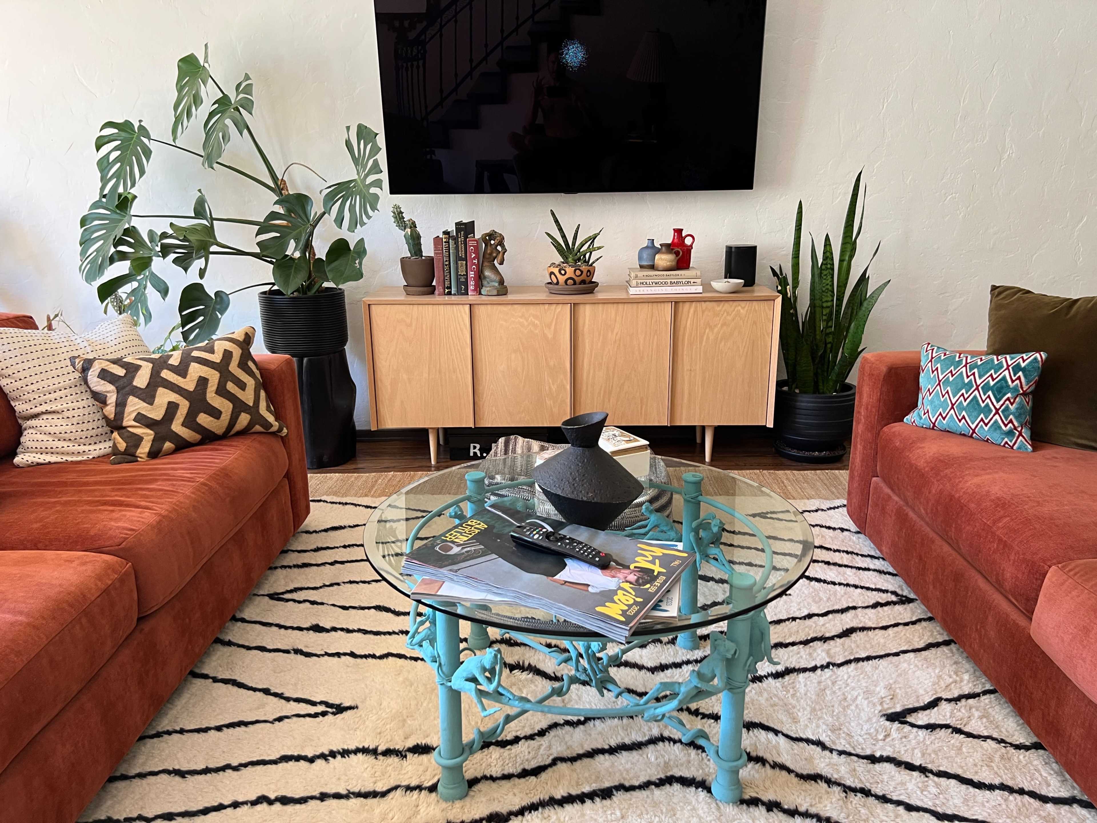 A living room features two orange sofas flanking a circular glass coffee table on a striped rug, with a television mounted on a light-colored wall.
