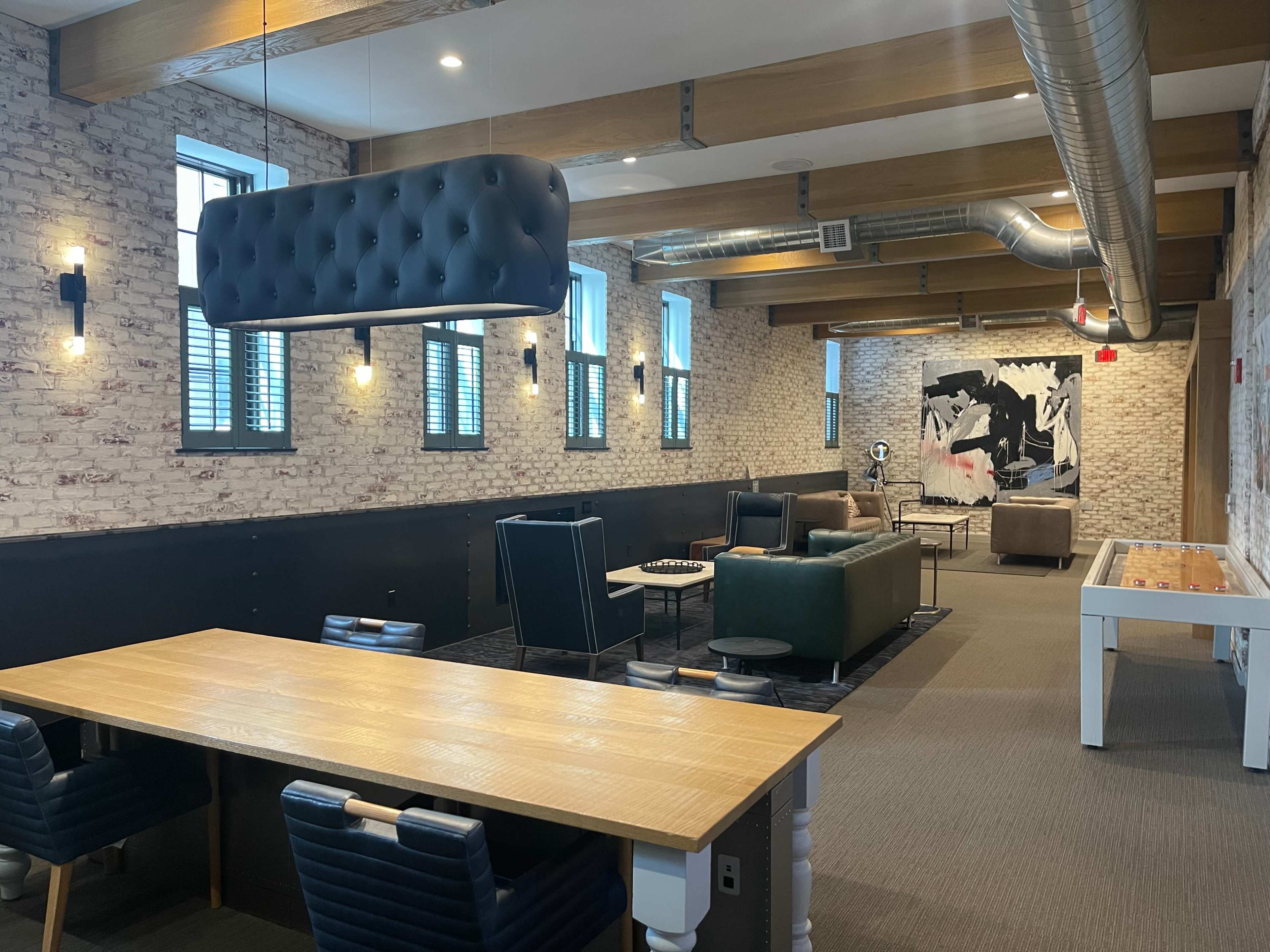 The image shows a spacious lounge area with exposed brick walls, seating arrangements including sofas and chairs, and a large wooden table.