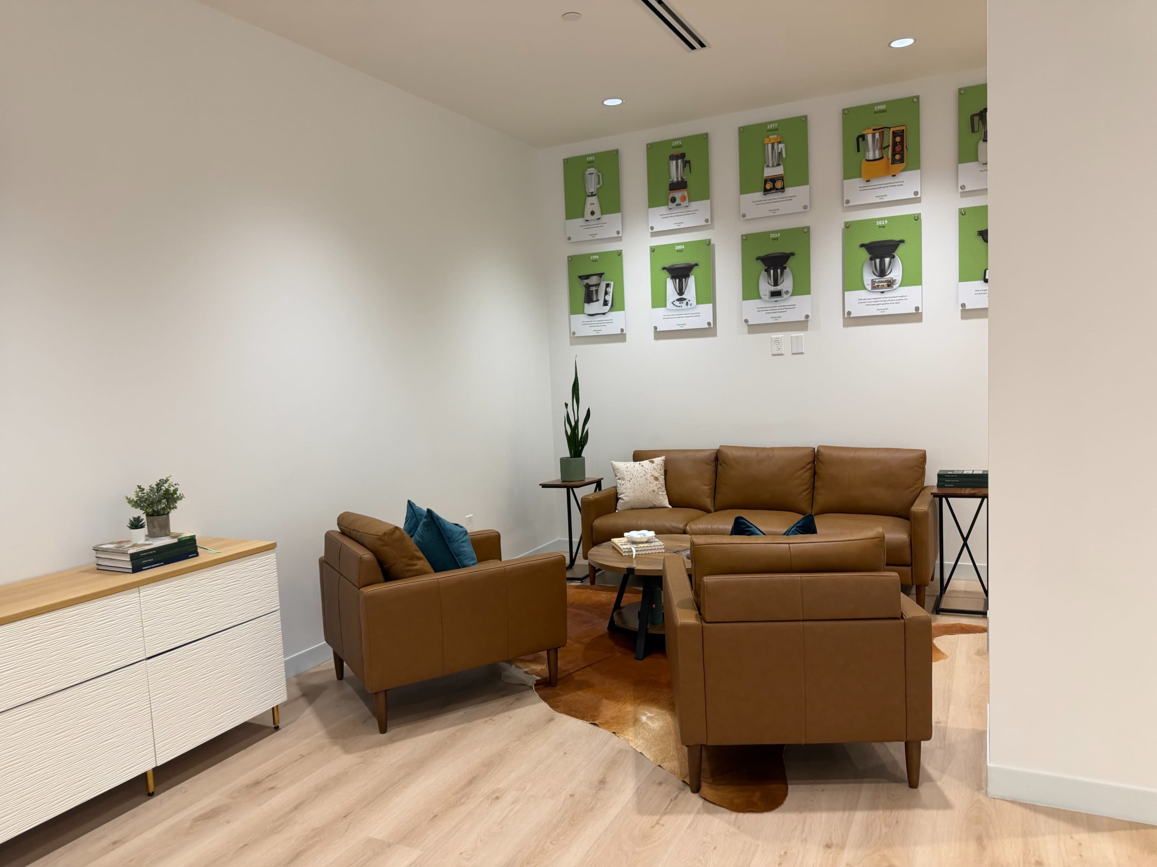 The image depicts a modern lounge area with brown leather seating, a wood side table, and several green and white wall displays featuring products.