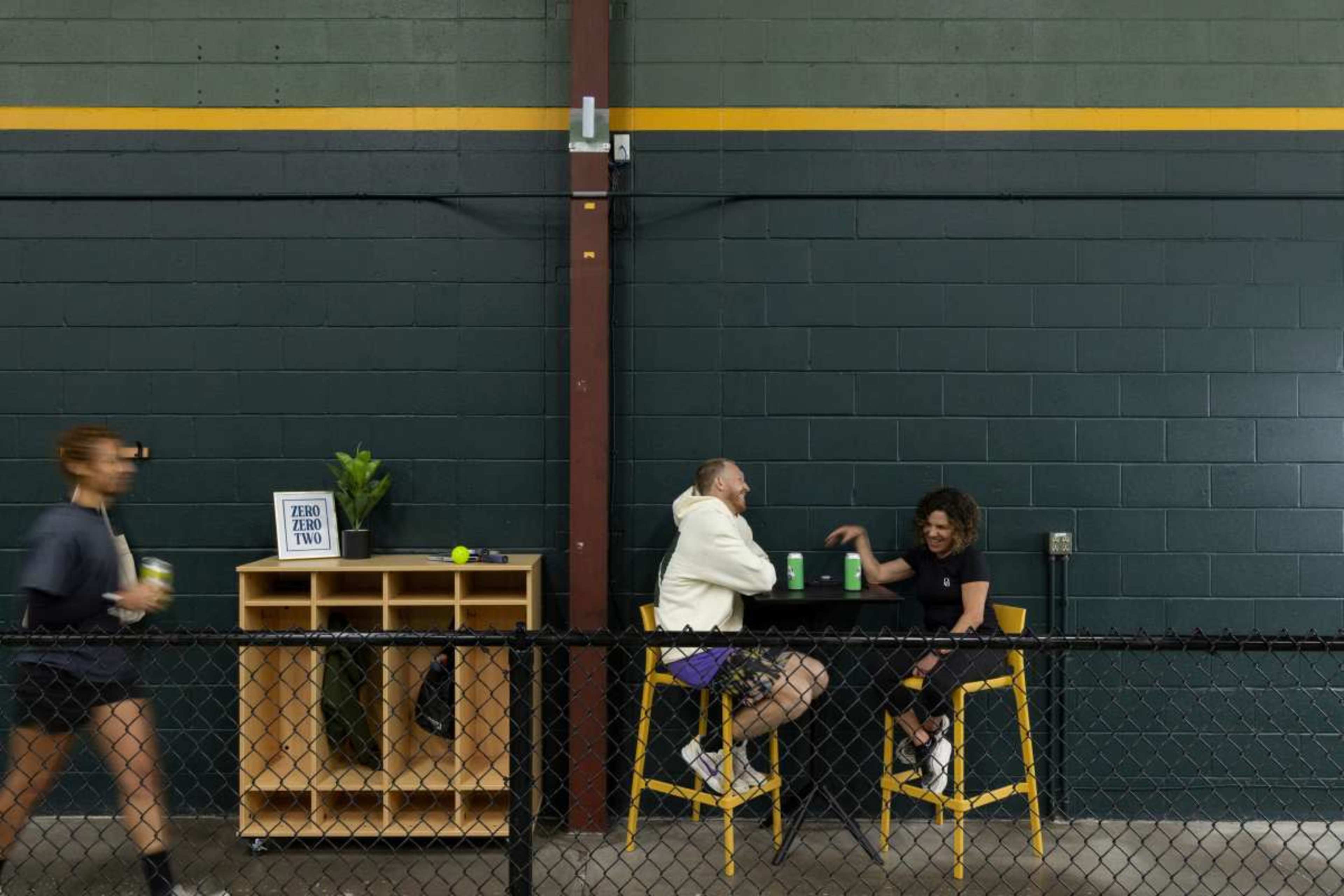 Two people sit at a high table engaged in conversation, while a third person walks by in the background.