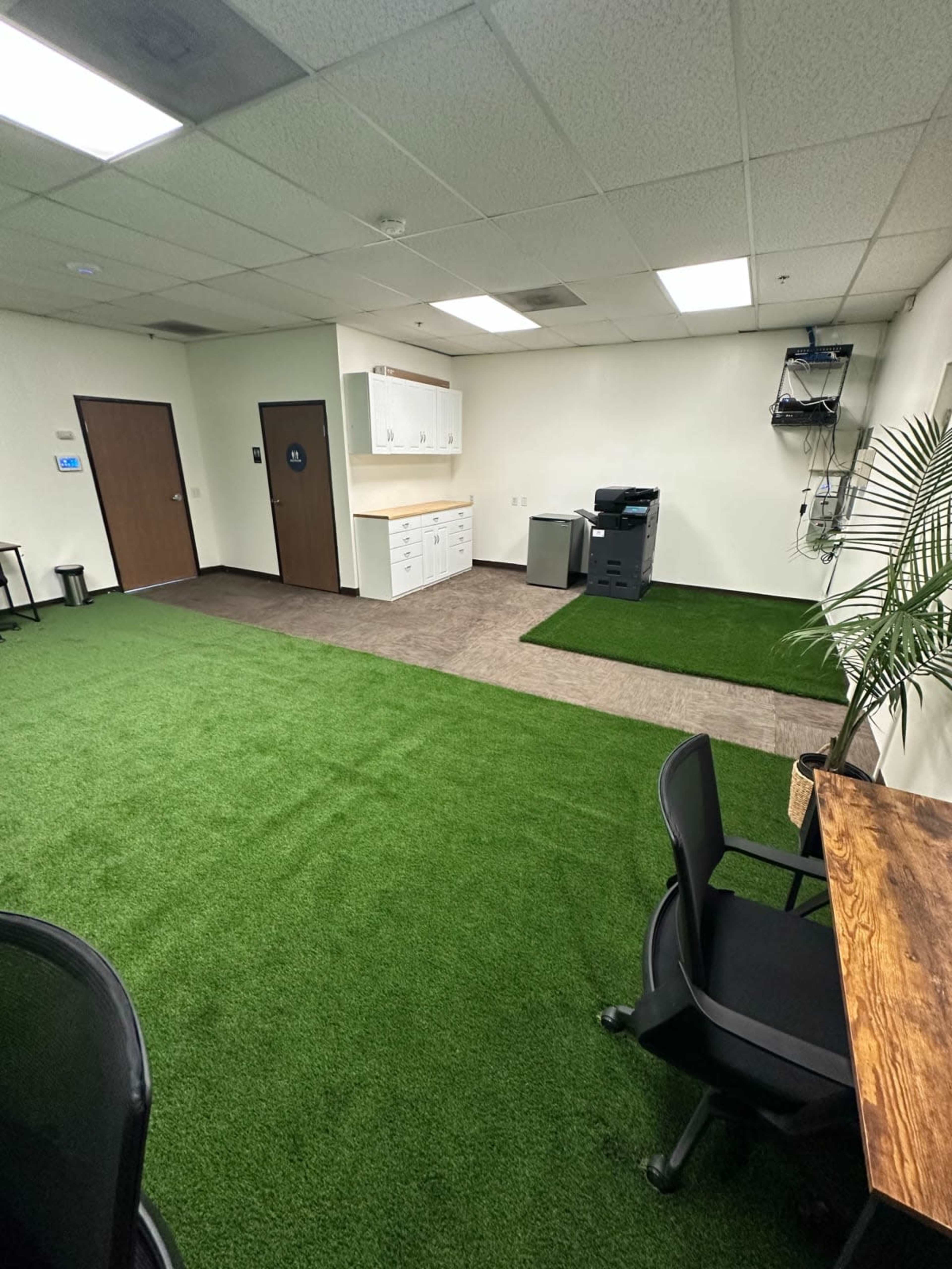 The image shows an office space featuring green carpet-like flooring, a kitchenette area with white cabinets, a small refrigerator, and a printer, along with a desk and chairs.
