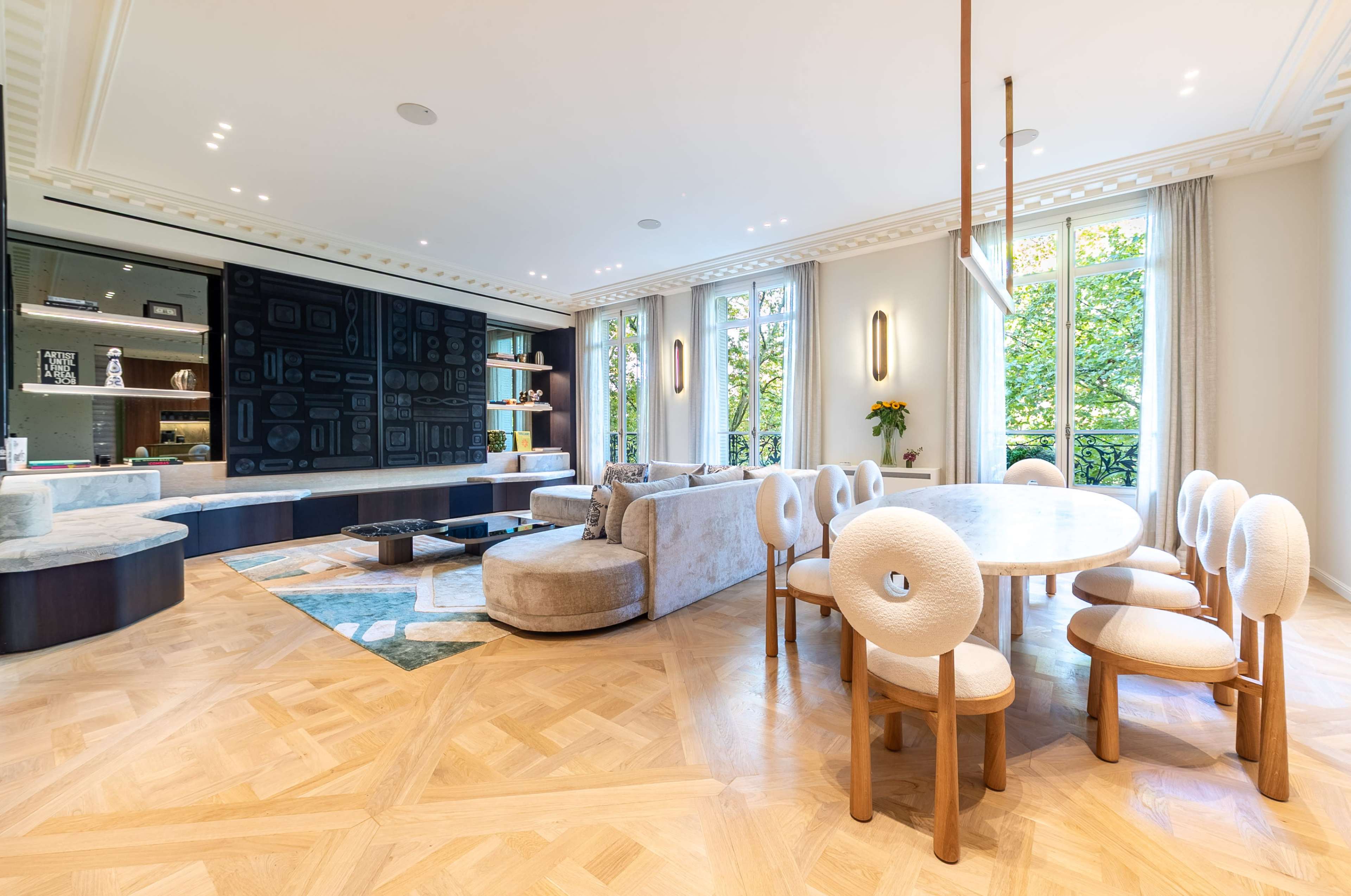 The image shows a modern living area with a sectional sofa, a circular dining table surrounded by upholstered chairs, and large windows allowing natural light in, all complemented by wooden flooring.