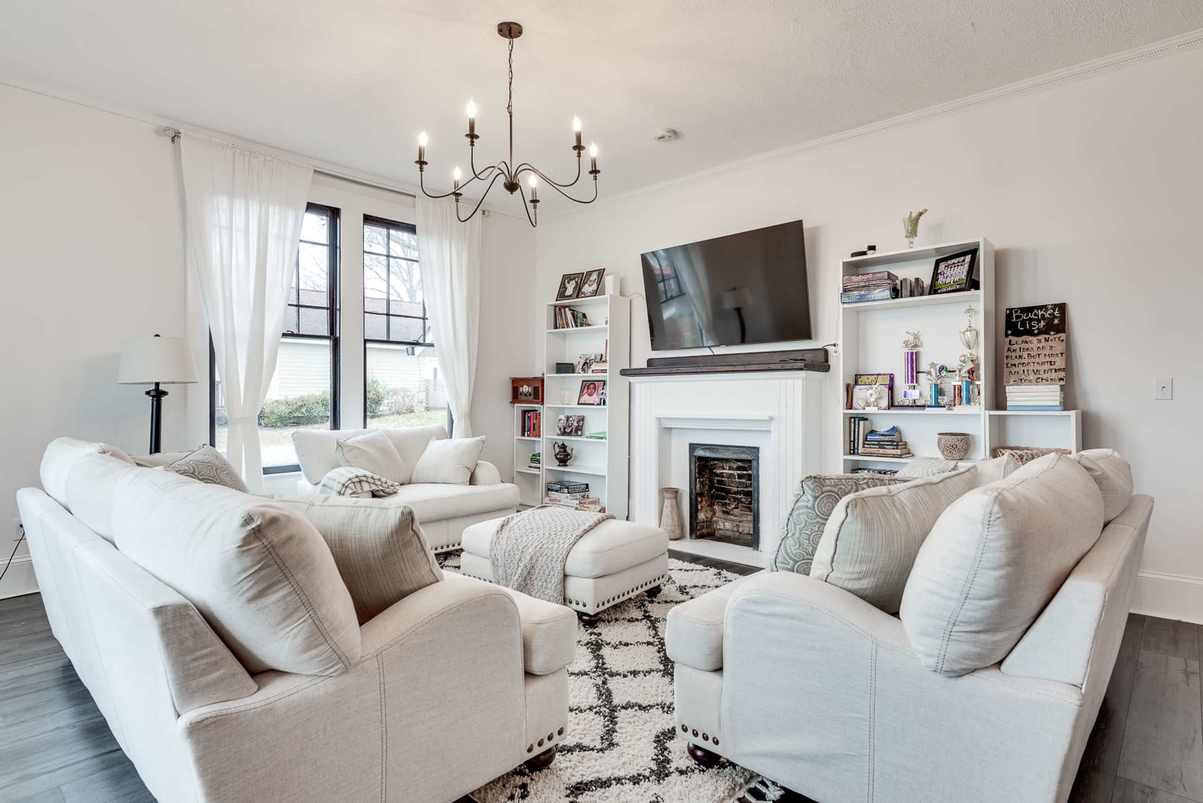 The living room features a chandelier, a large television mounted above a fireplace, and white sofas arranged around a patterned rug.