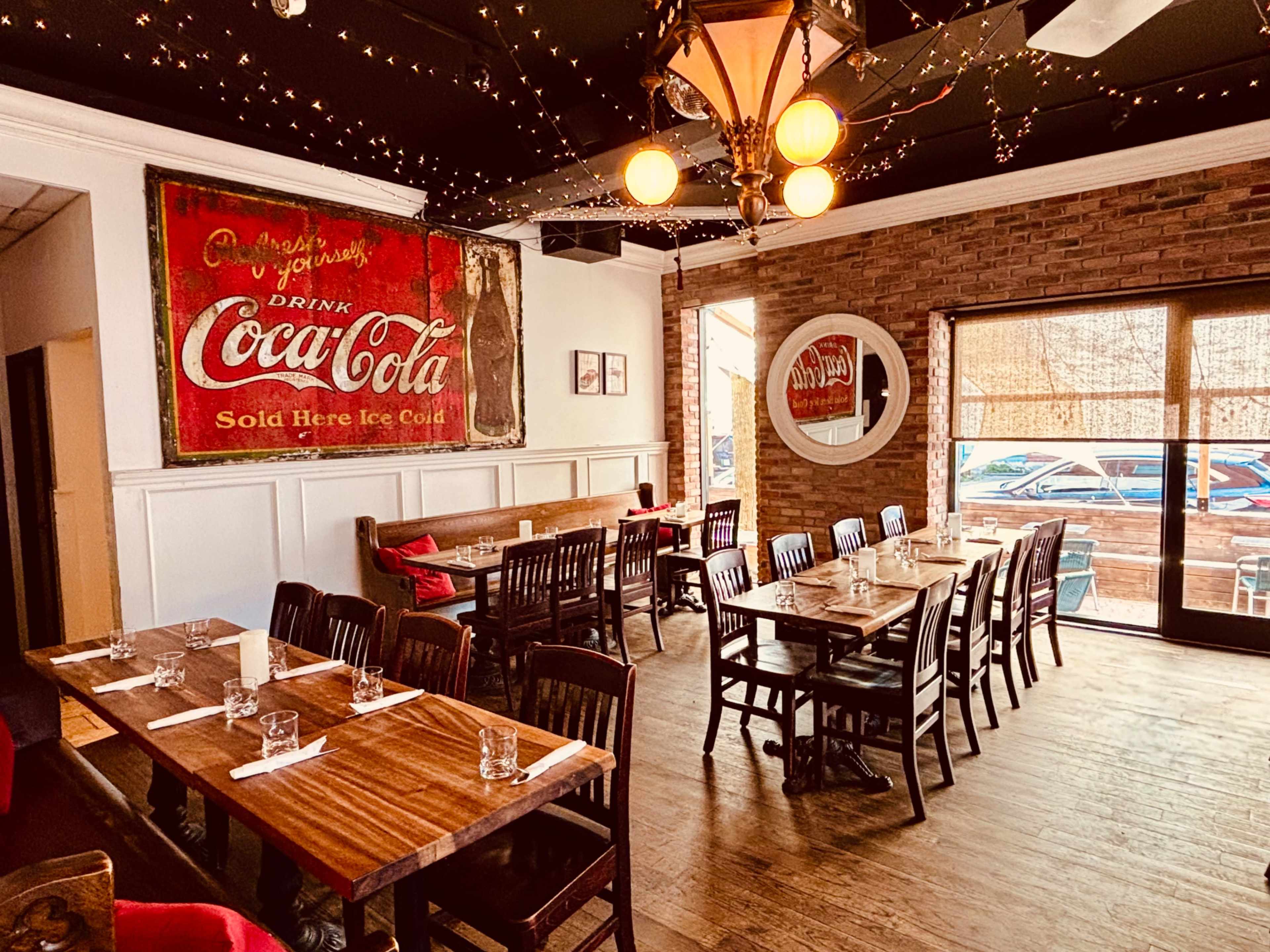 The image shows a restaurant interior with wooden tables and chairs, a large vintage Coca-Cola sign on the wall, and string lights hanging from the ceiling.