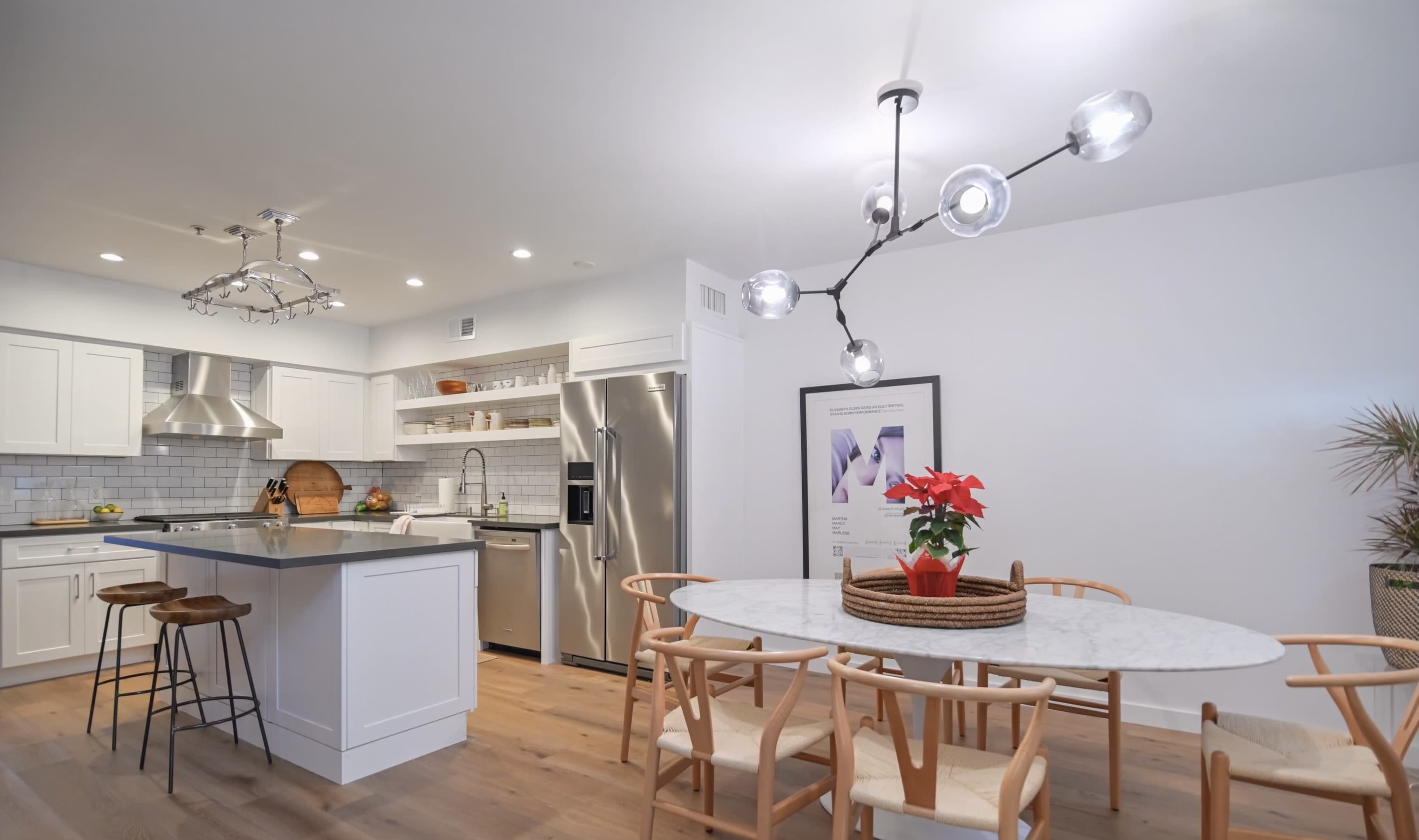 The image shows a modern kitchen and dining area featuring white cabinetry, a stainless steel refrigerator, a marble dining table with wooden chairs, and a decorative light fixture overhead.
