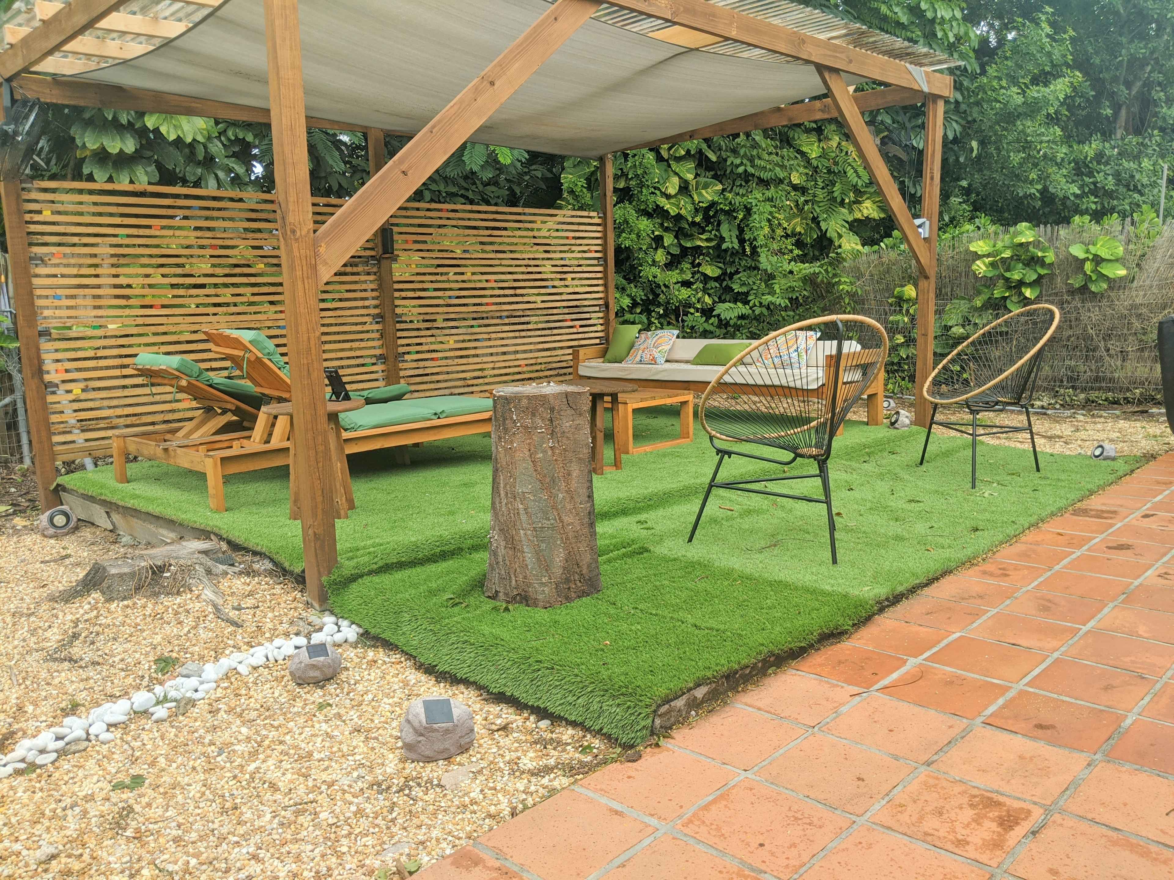 A shaded outdoor seating area features a wooden pergola, two metal chairs, a lounge chair, and a tree stump on artificial grass with gravel and stone accents.