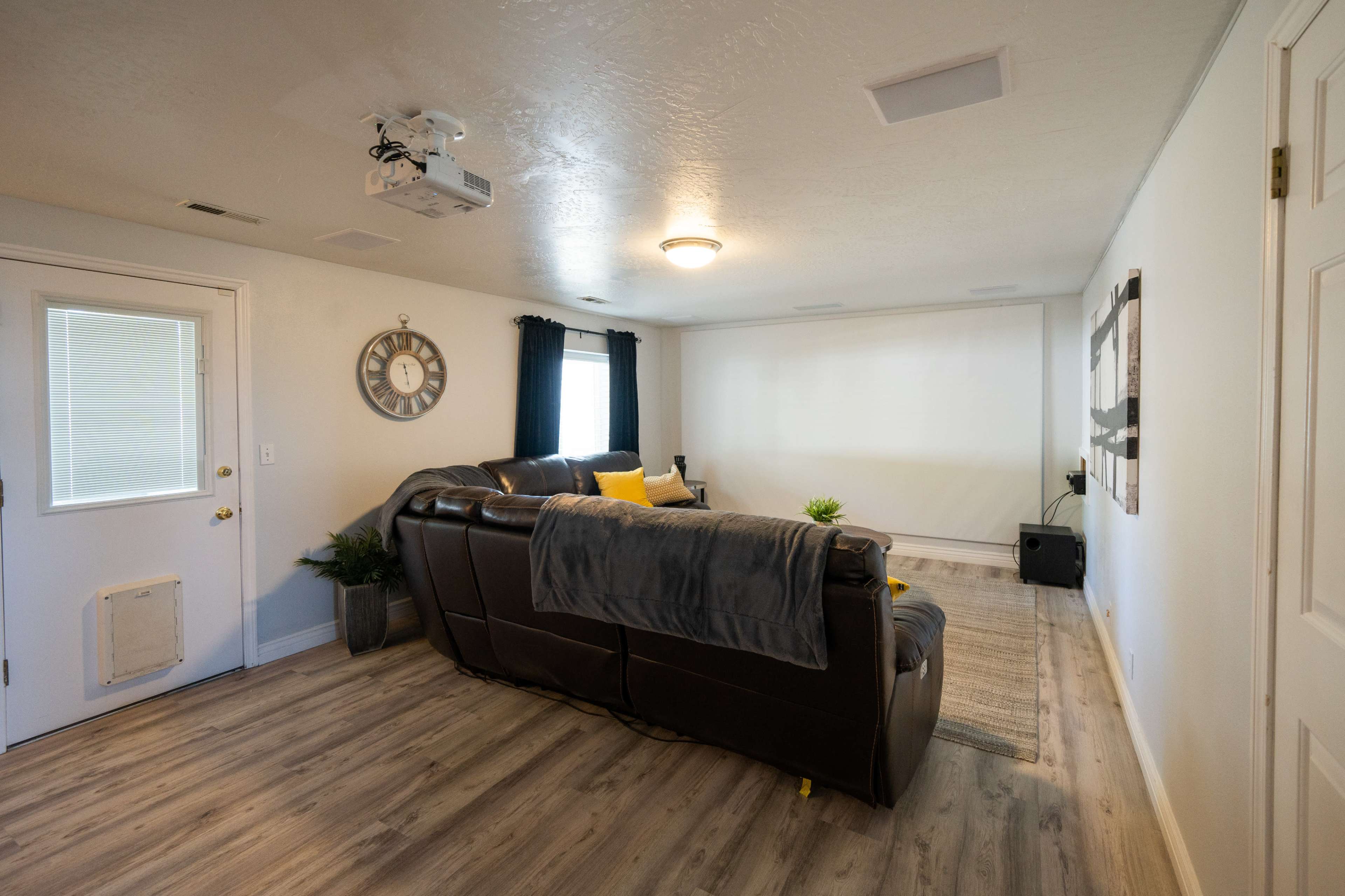 A living room features a large sectional couch, a wall-mounted projector setup, and a door leading outside.