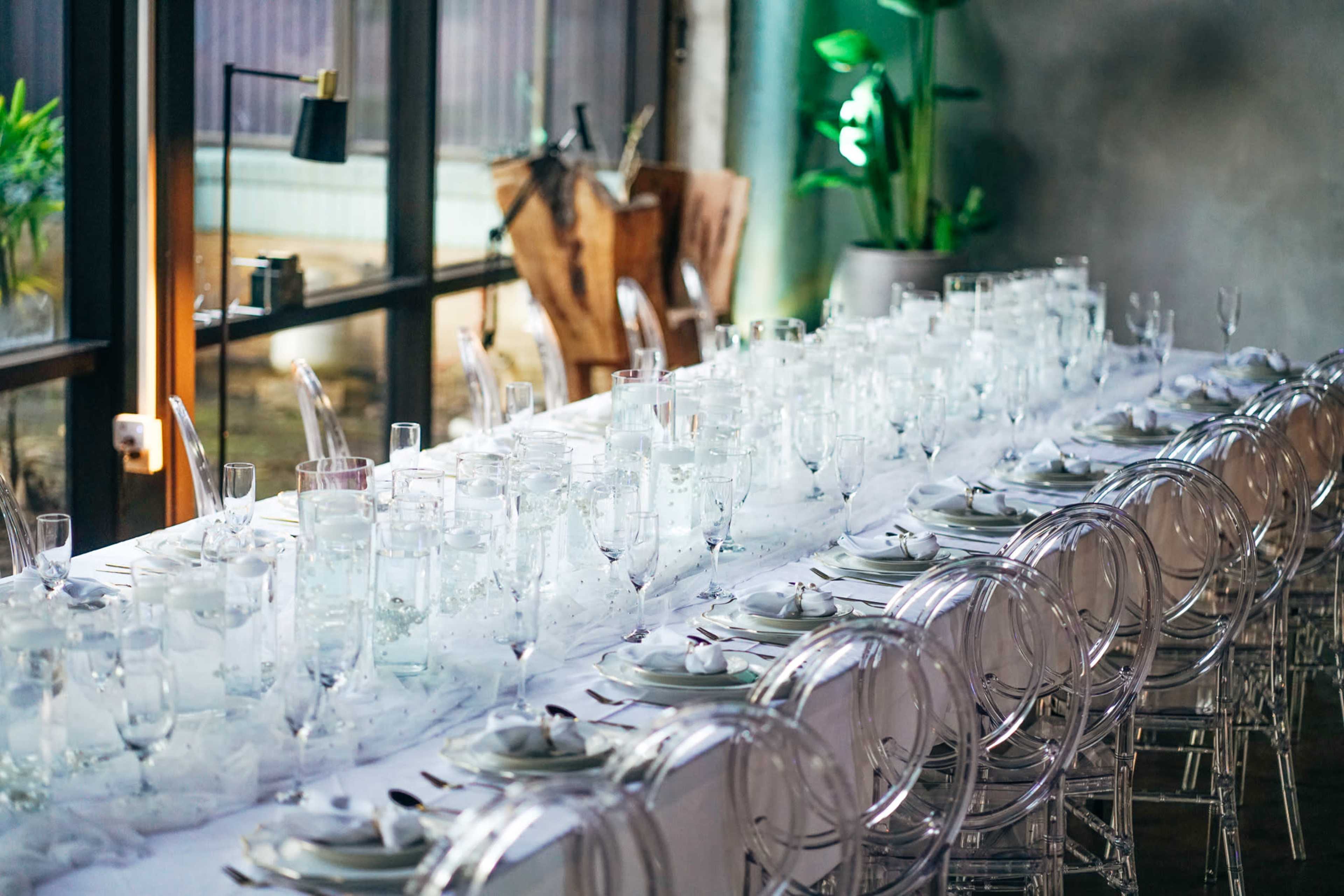 A long dining table is set with clear glassware and white tablecloths, surrounded by transparent chairs in a well-lit indoor space.