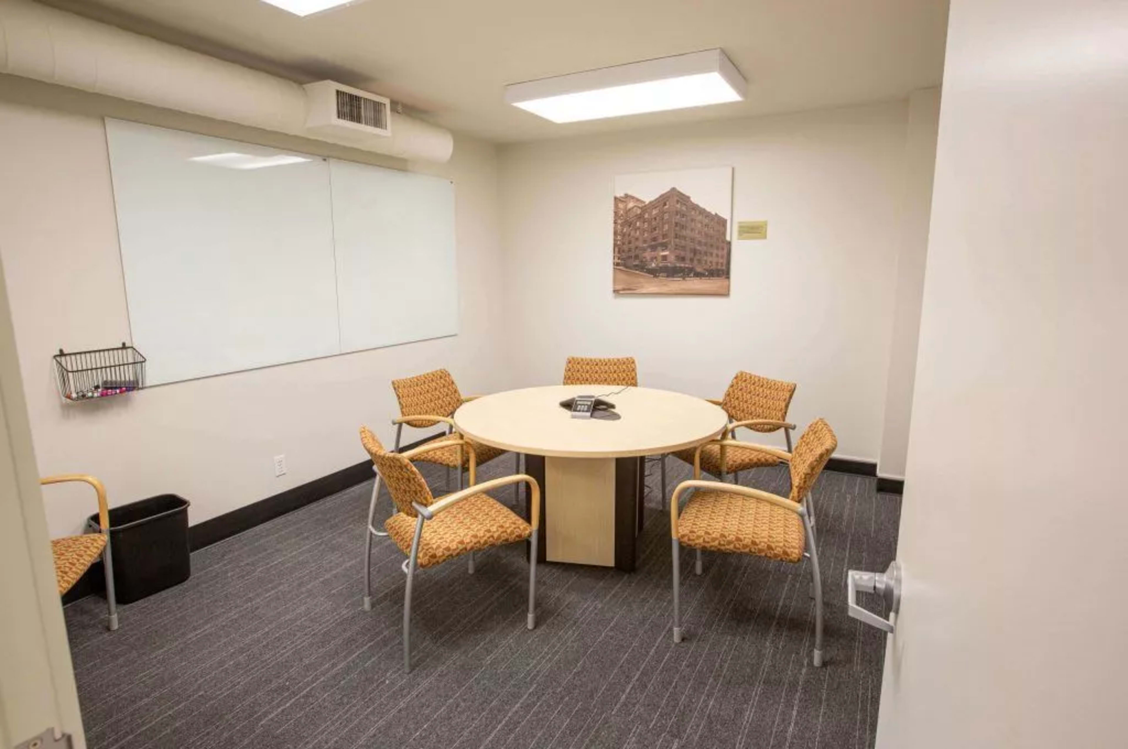 A small meeting room with a circular table surrounded by four orange chairs, a whiteboard on the wall, and a wastebasket nearby.