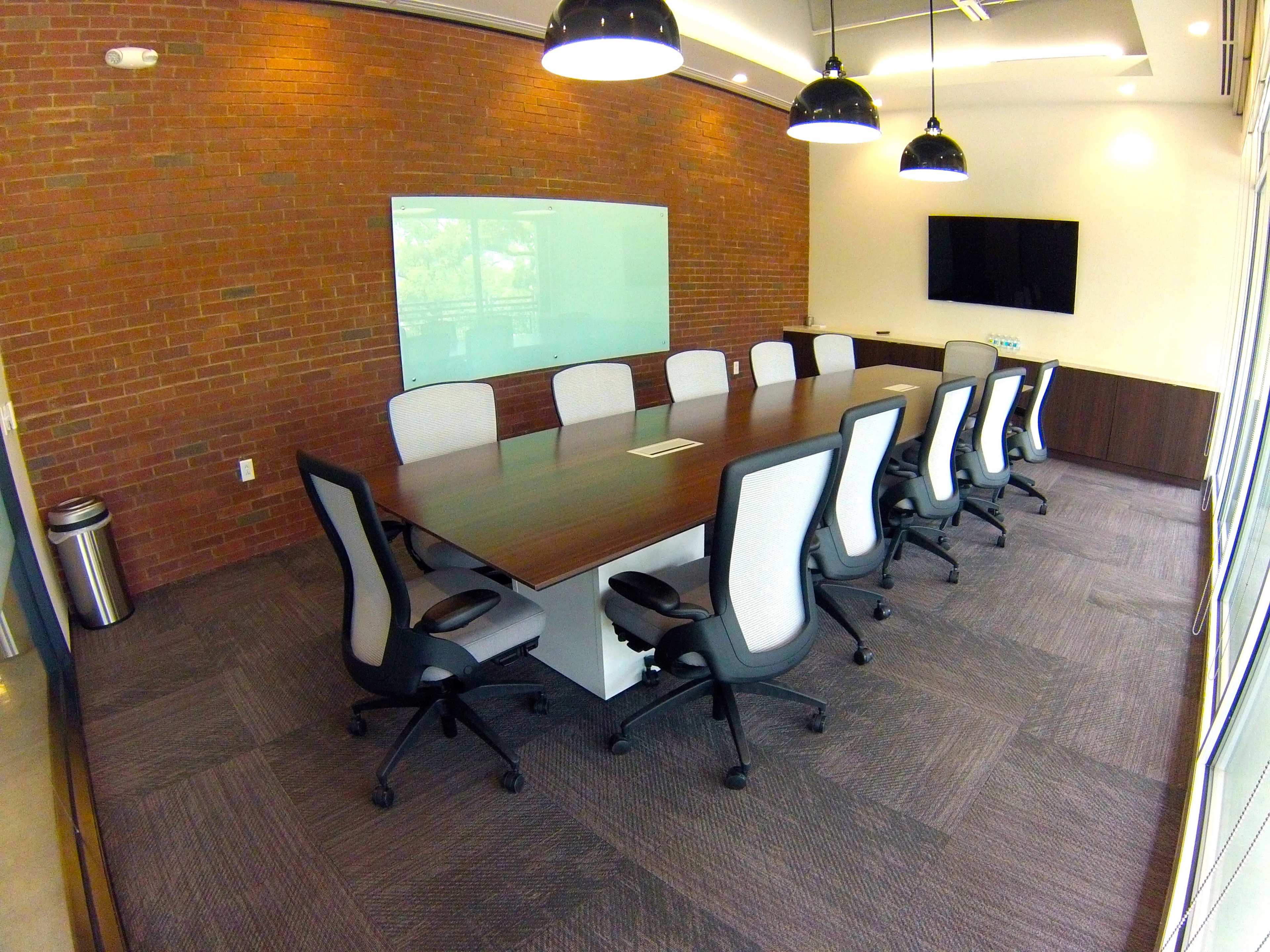A modern conference room with a long wooden table surrounded by ergonomic chairs, a large screen on the wall, and a brick accent wall.