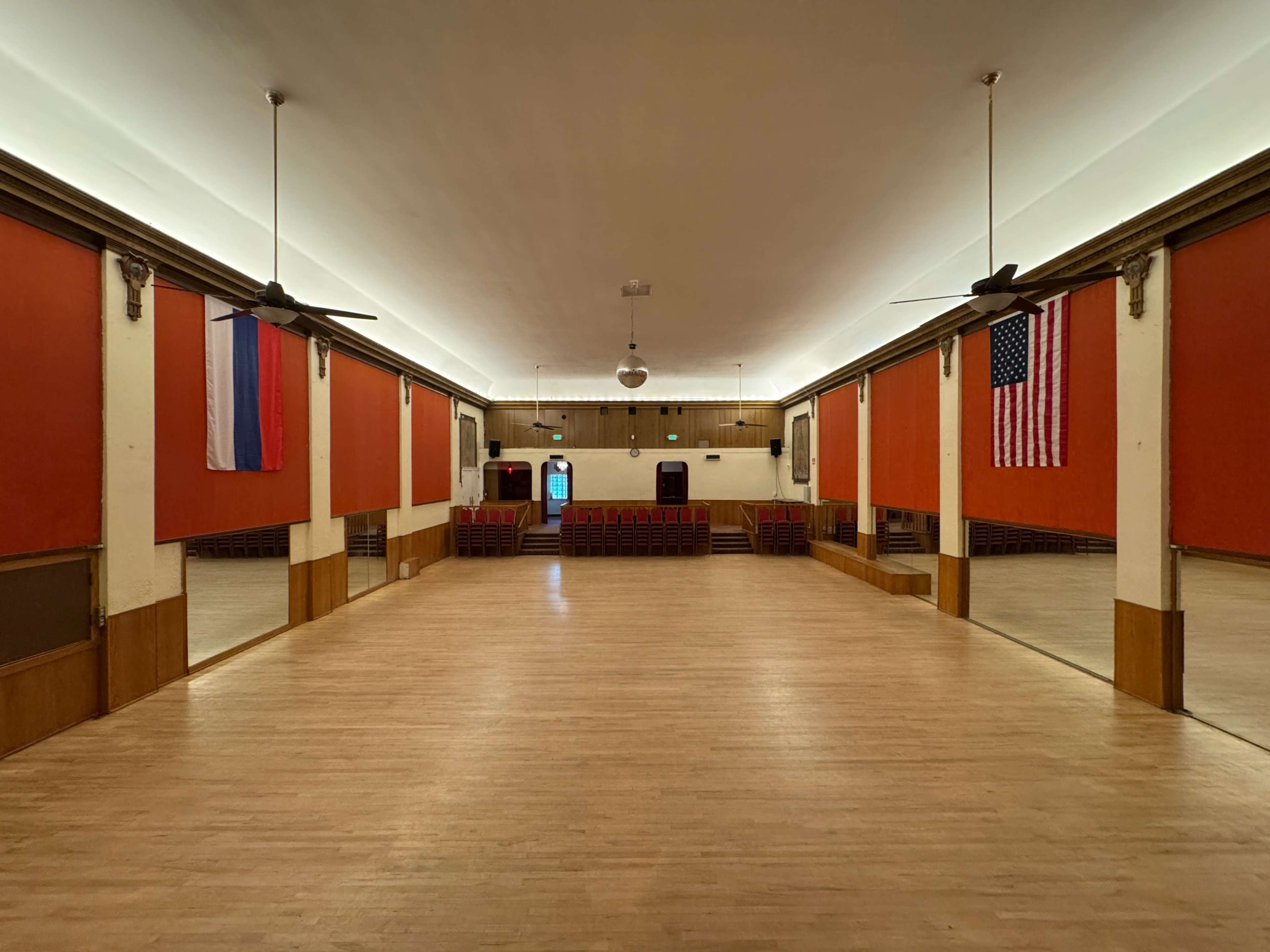 The image shows a spacious, empty hall with wooden flooring, adorned with walls featuring red panels and a Russian and an American flag hanging at either end.