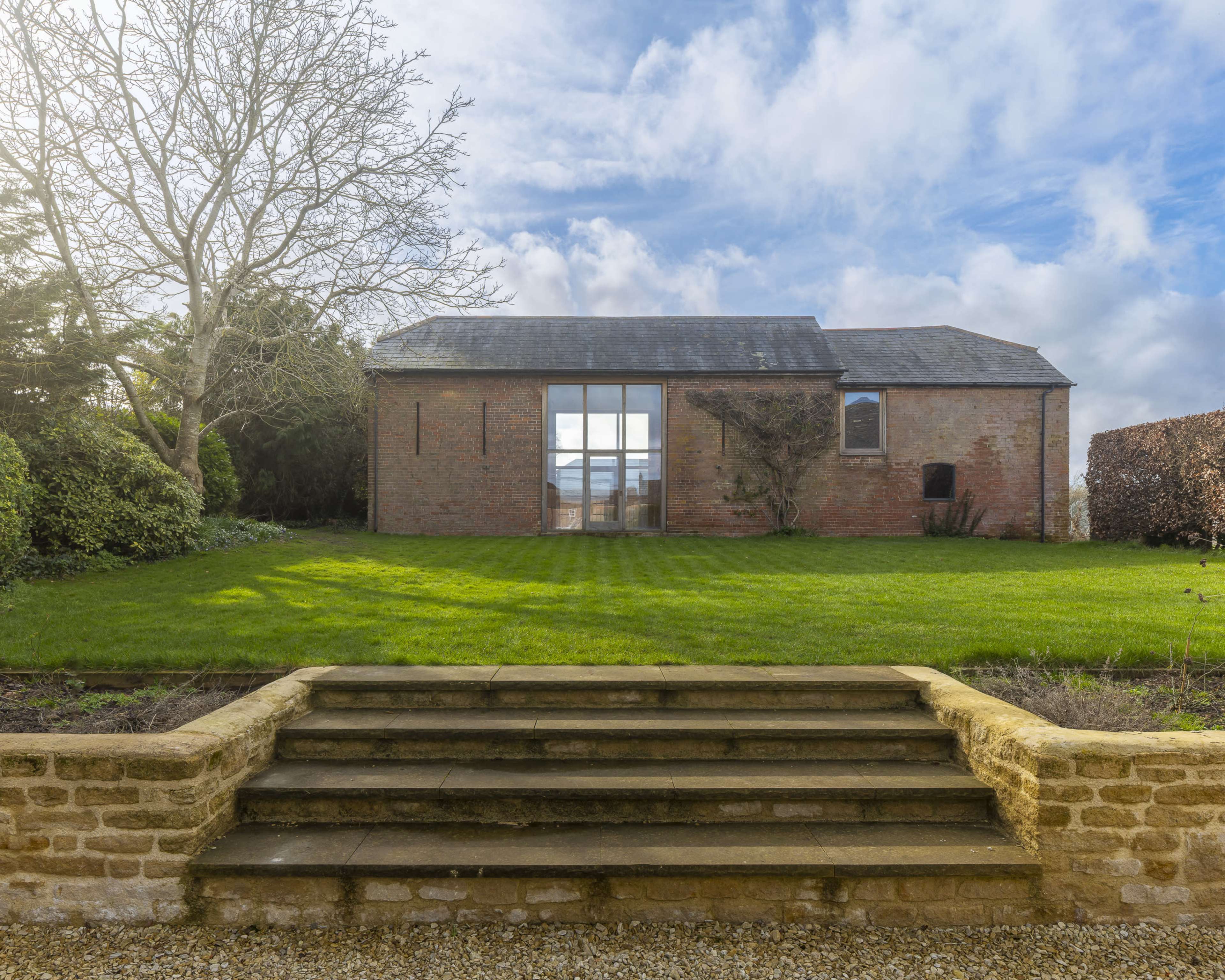 A brick building with large glass doors is set against a green lawn and a cloudy sky, with stone steps leading up to it.