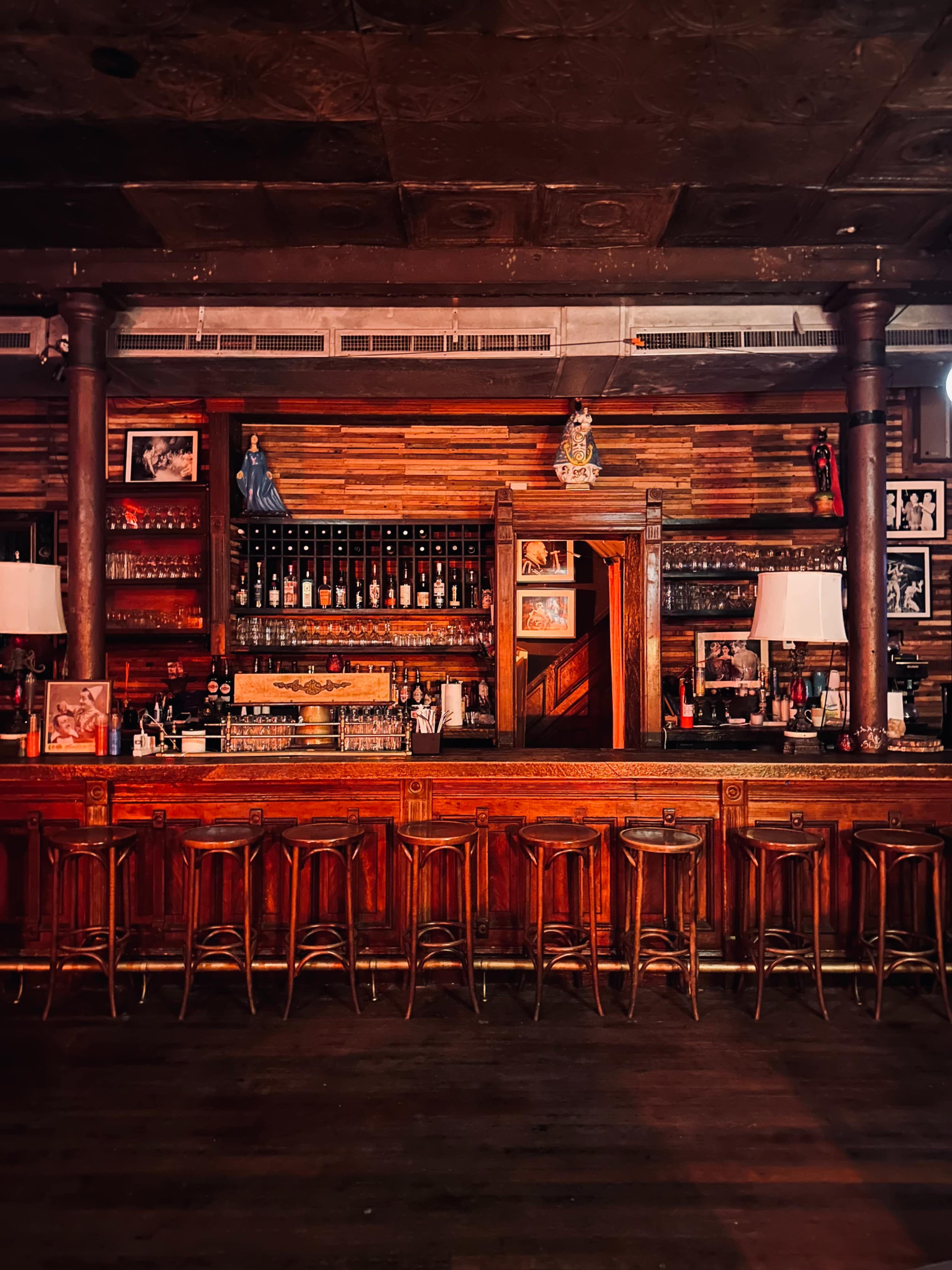 The image shows a rustic bar with a long wooden counter, surrounded by high-backed stools, and adorned with bottles and framed photographs on the walls.
