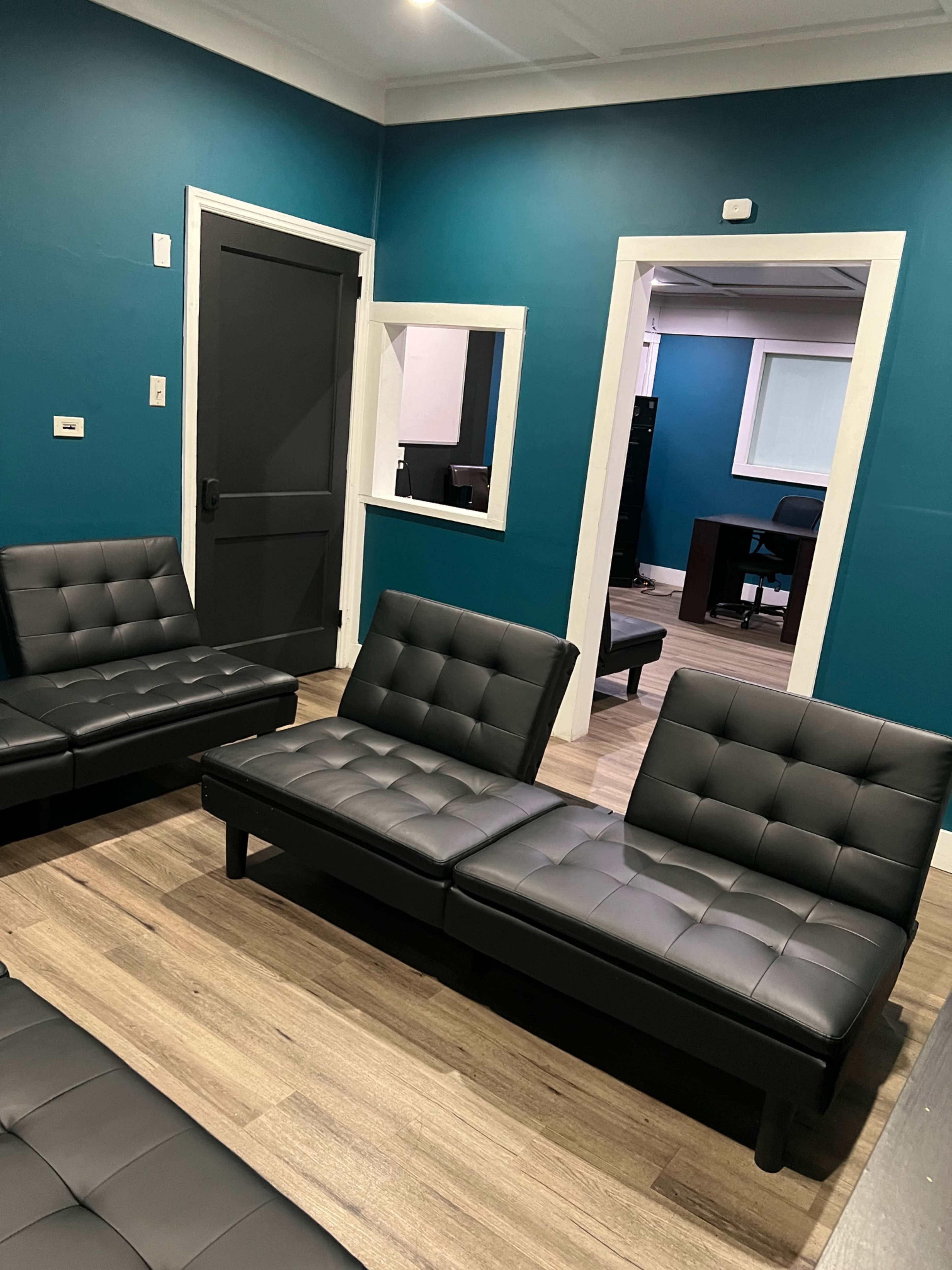 The image shows a waiting area with black lounge chairs, teal walls, and a doorway leading to a separate room with a desk and chair visible.