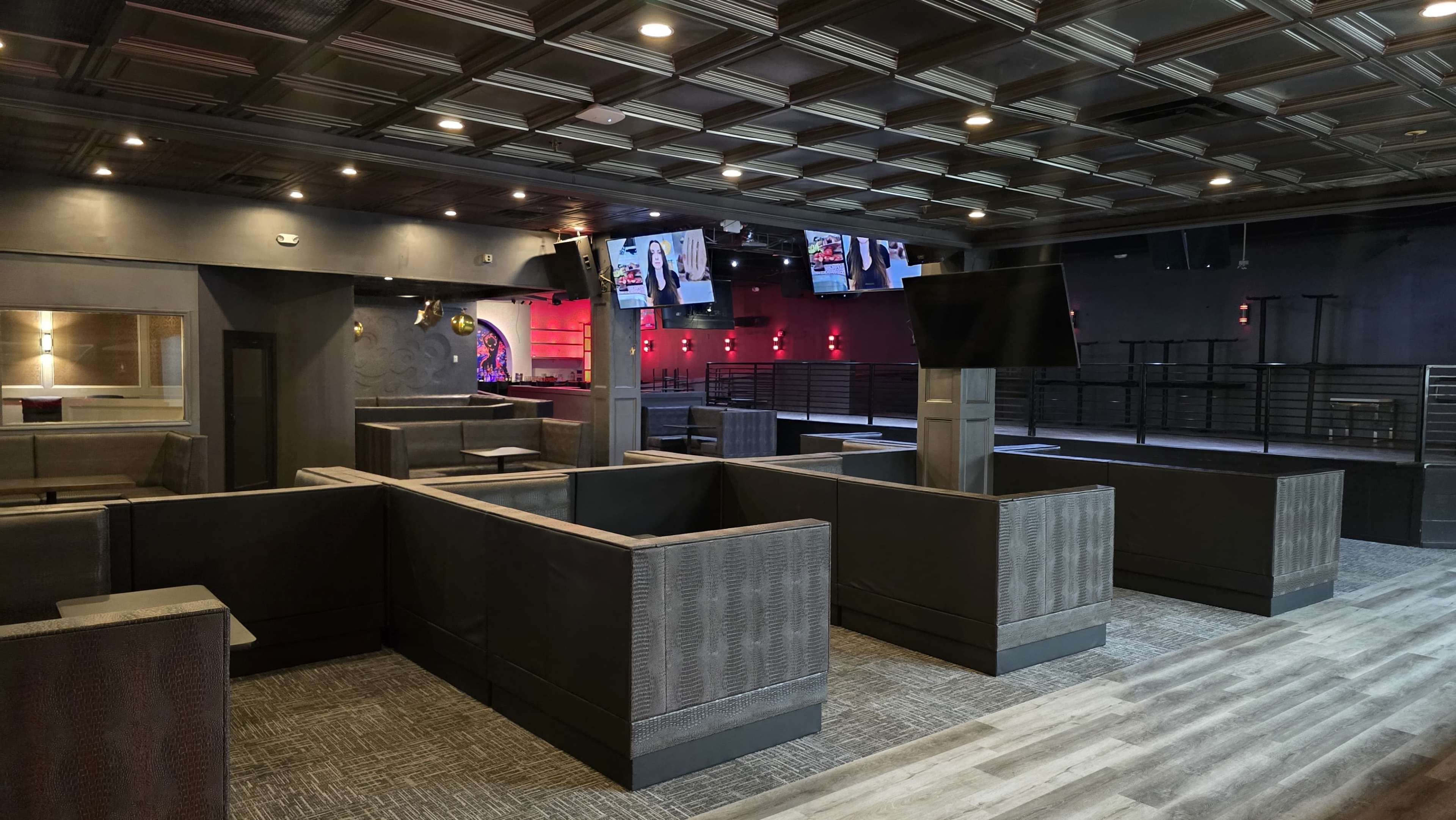 The image shows an empty lounge or bar area with dark seating arrangements, dim lighting, and multiple screens displaying content.