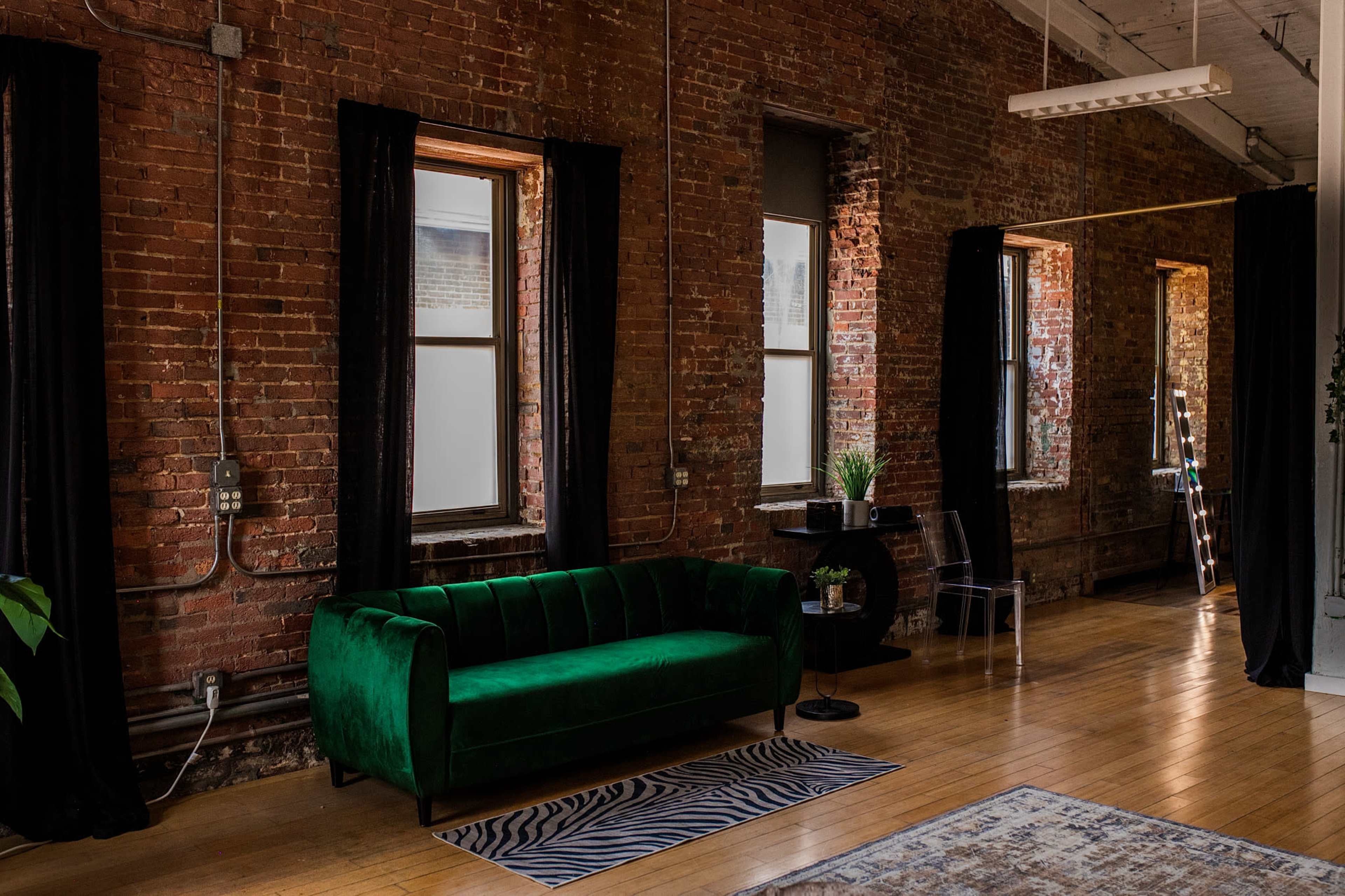 The image shows a stylish interior with exposed brick walls, large windows draped in black curtains, a green velvet couch, and a zebra-patterned rug on a wooden floor.
