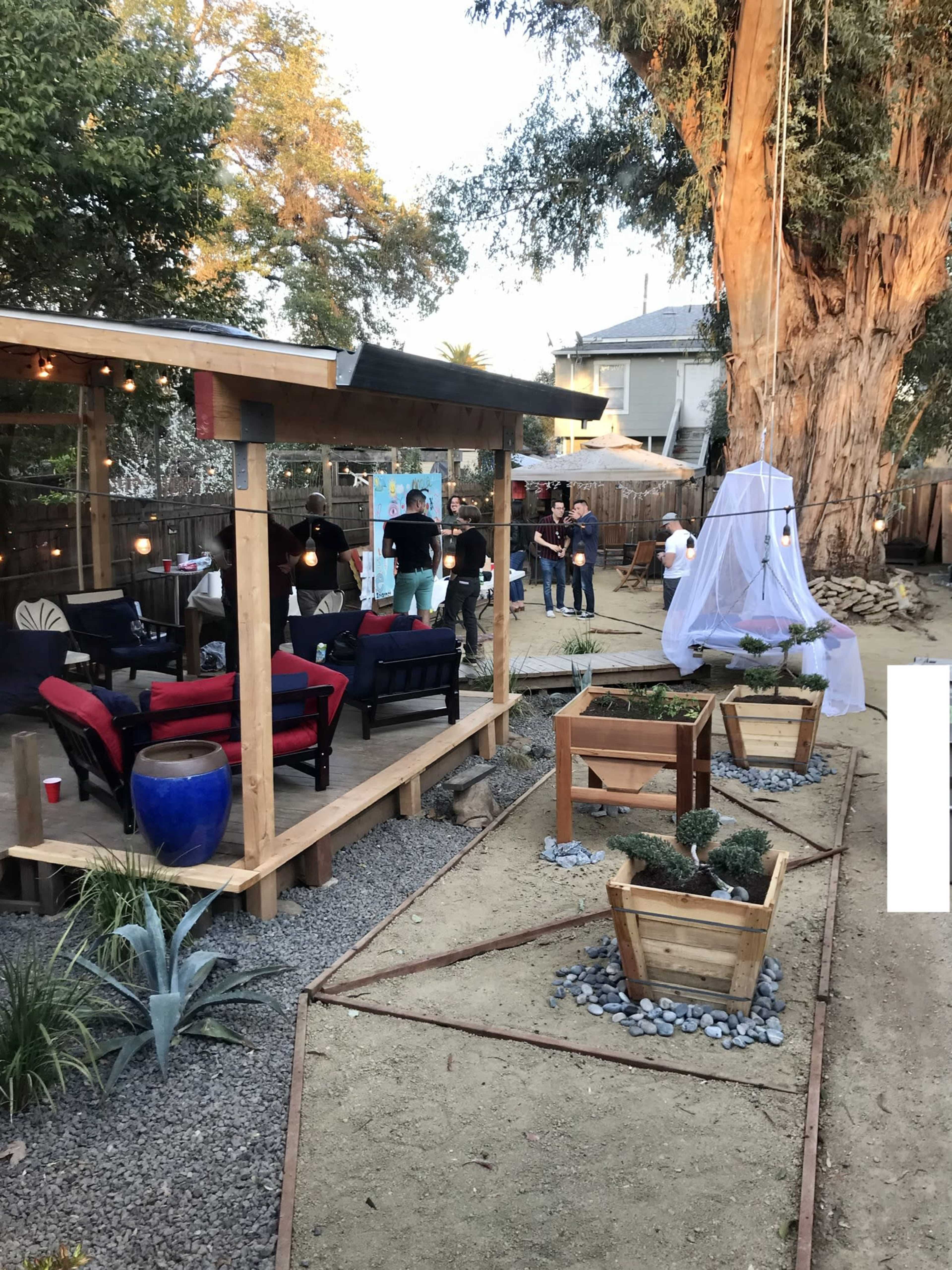 The image shows a backyard with a wooden patio area, several people gathered, and garden beds with small plants.