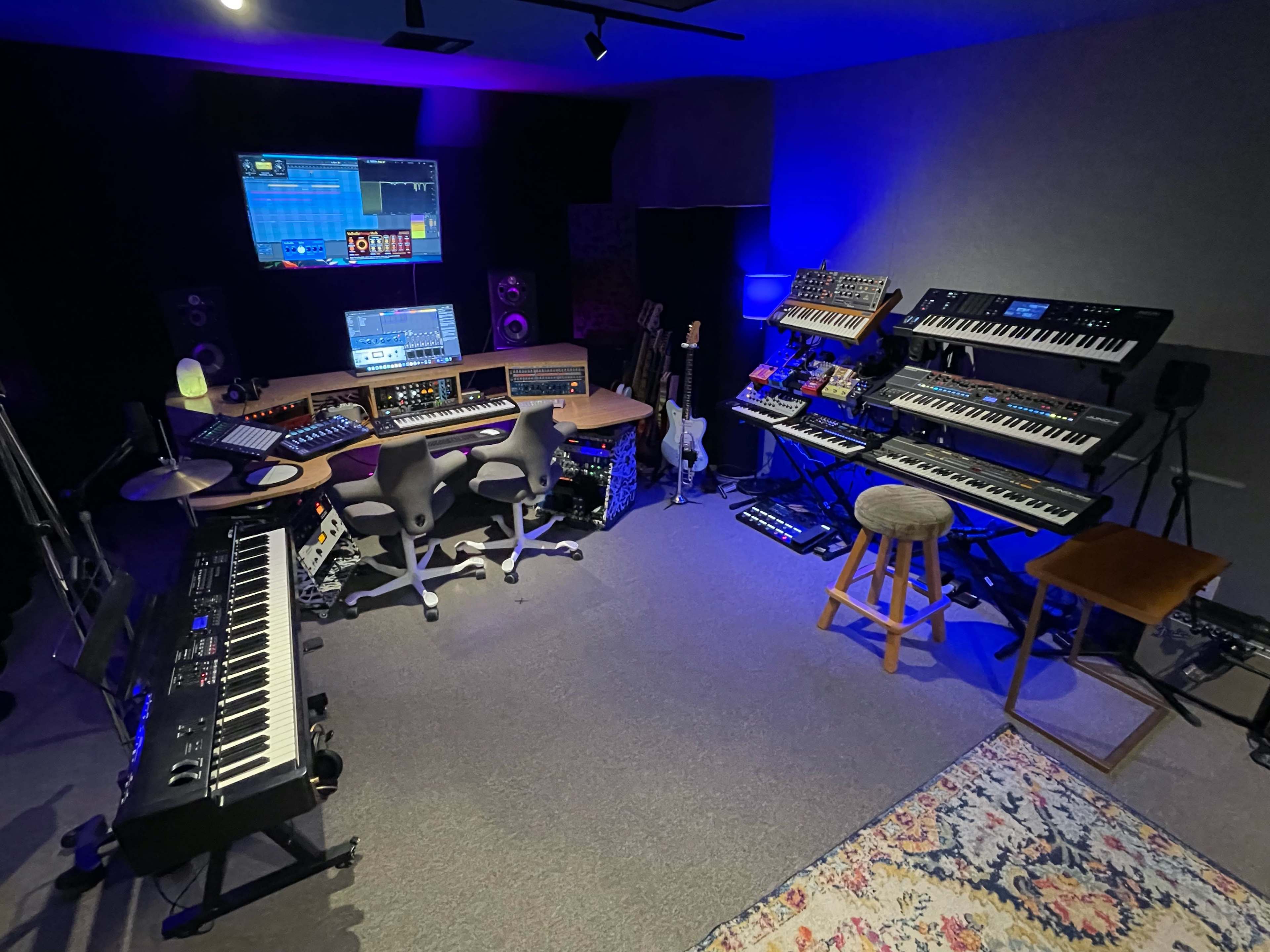 A music studio filled with various synthesizers, keyboards, and musical equipment arranged around a central workstation.