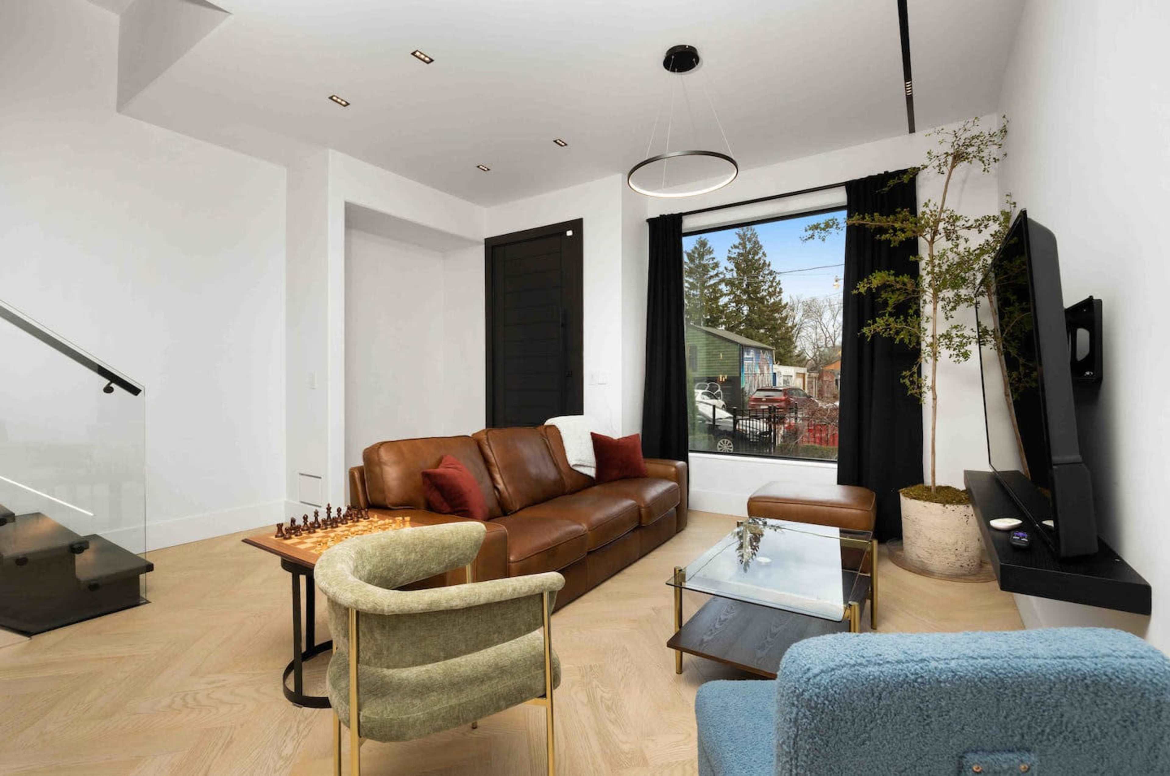 The image shows a modern living room featuring a brown leather sofa, a glass coffee table, a TV mounted on the wall, and a large window with indoor plants beside it.