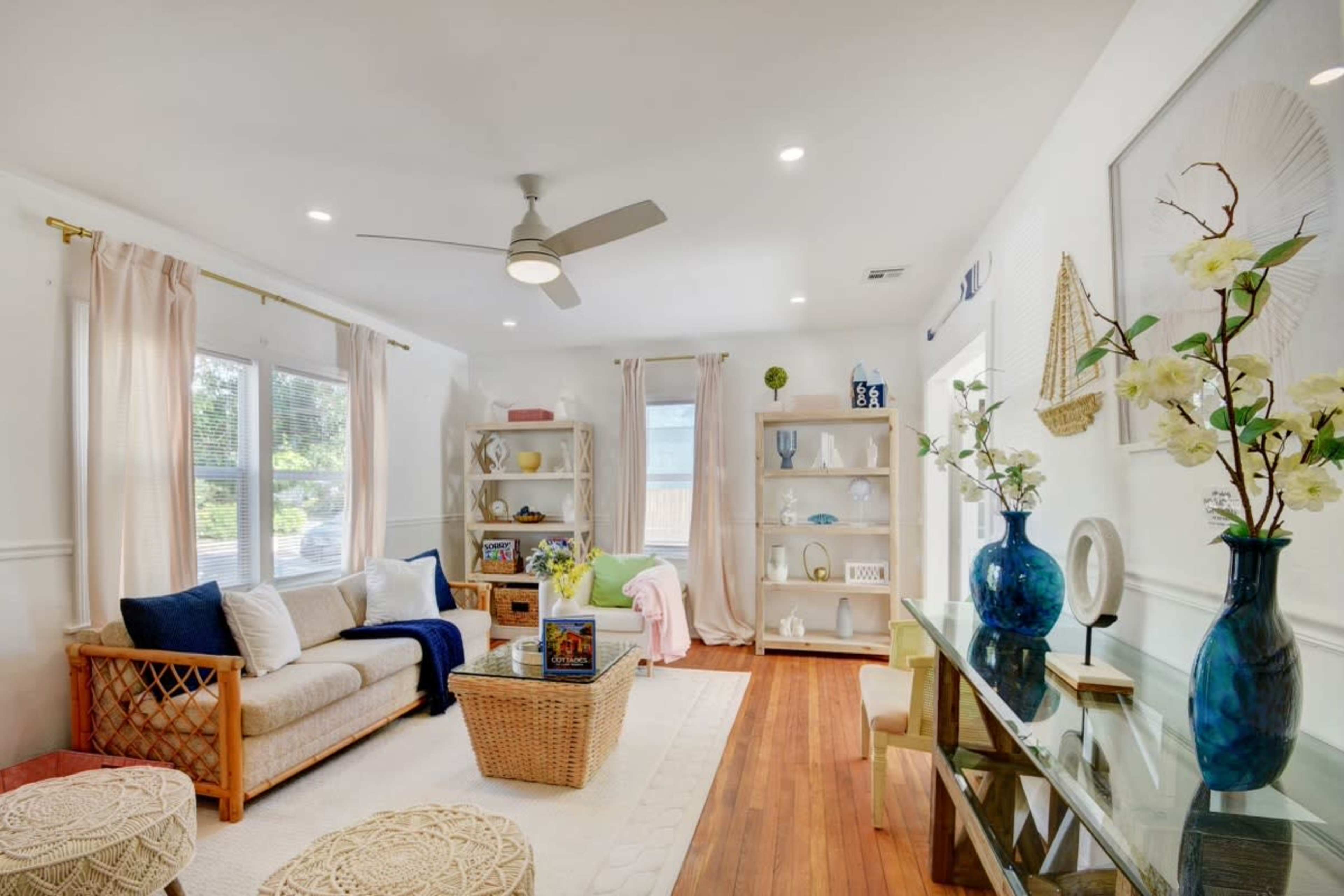 A bright living room featuring a sofa, a coffee table, and decorative shelves filled with plants and books, complemented by a ceiling fan and large windows.