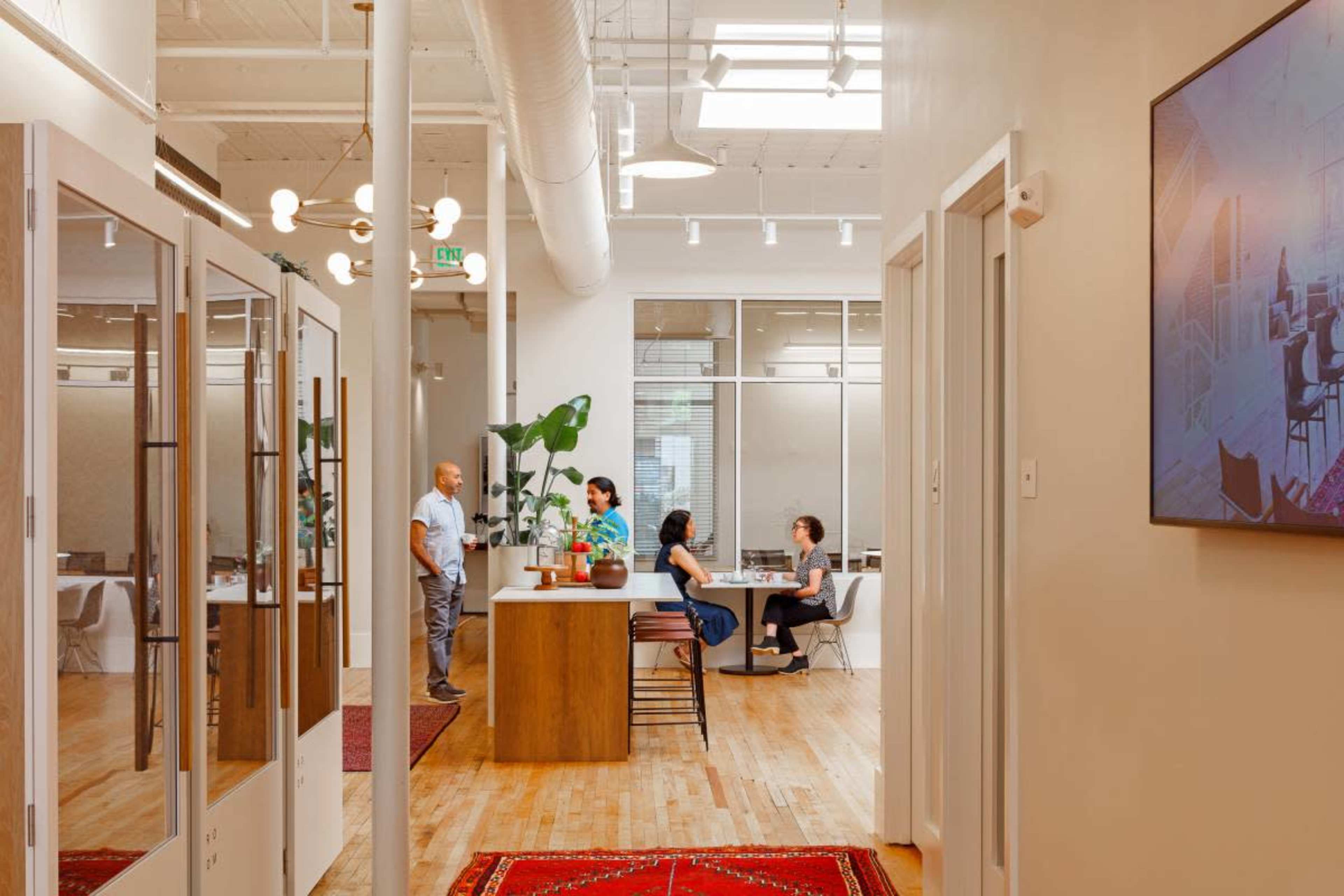 The image depicts a modern office interior featuring a communal workspace with a kitchenette and several individuals engaged in conversation.