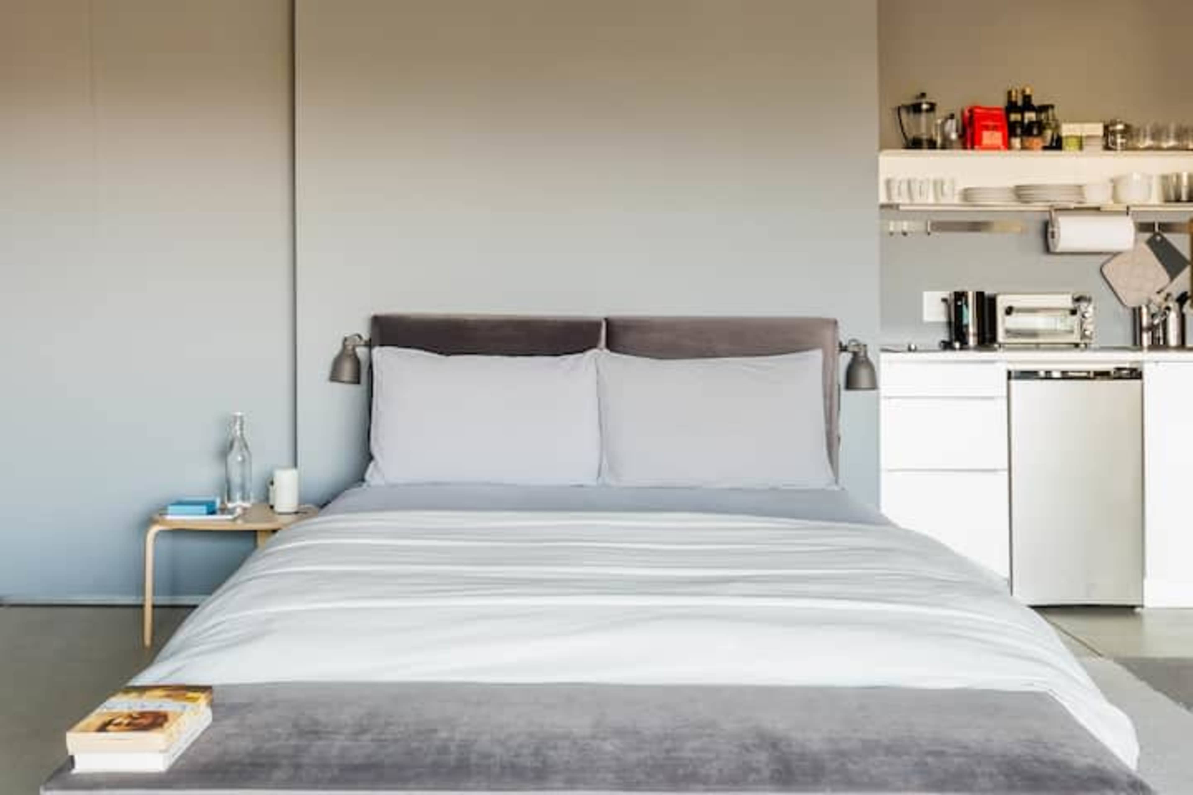 A minimalistic bedroom with a bed, two gray pillows, and a nightstand beside it, facing a kitchenette with various appliances and containers.