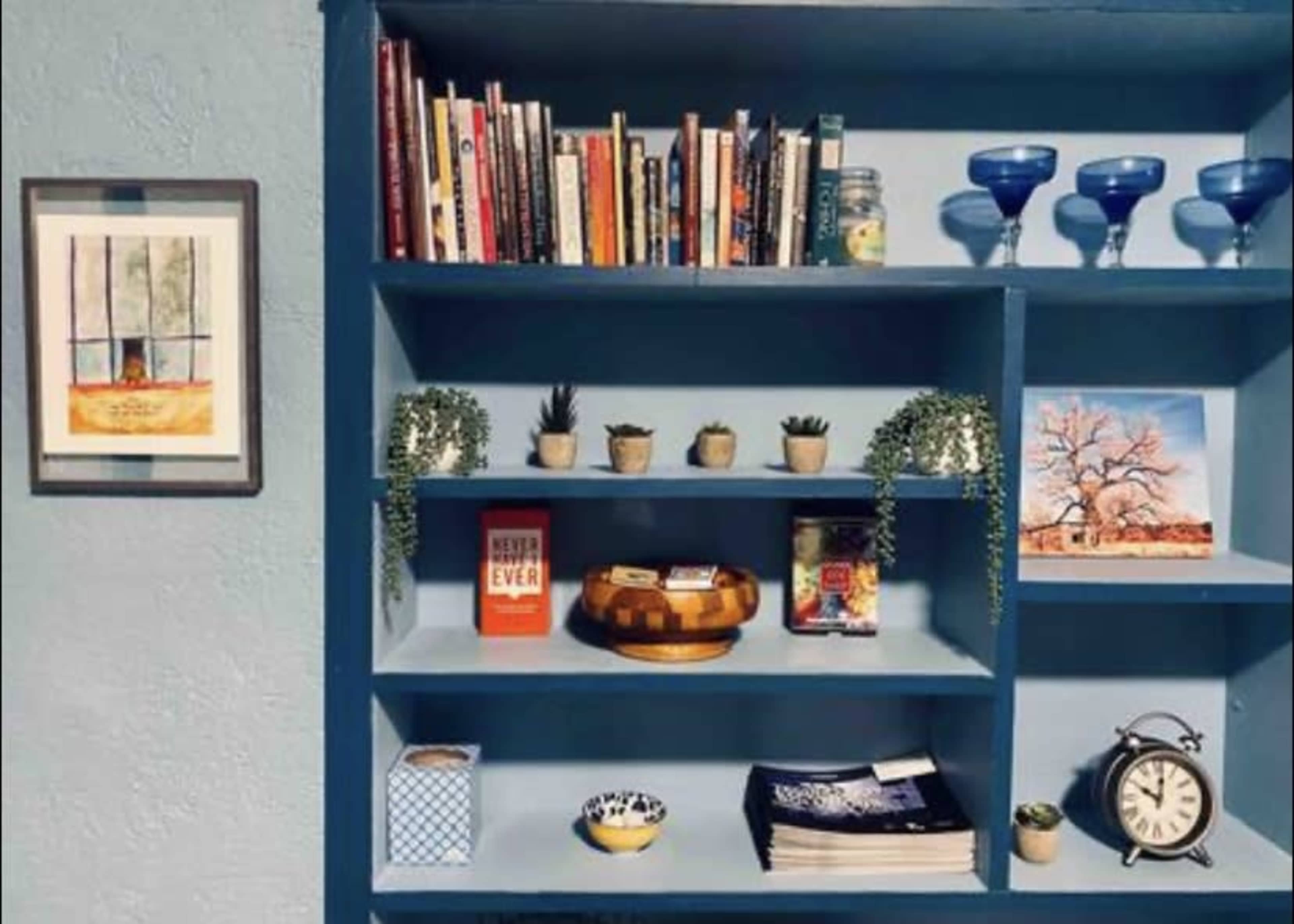 A blue bookshelf displays various books, decorative pots with plants, and framed artwork alongside a clock and other items.