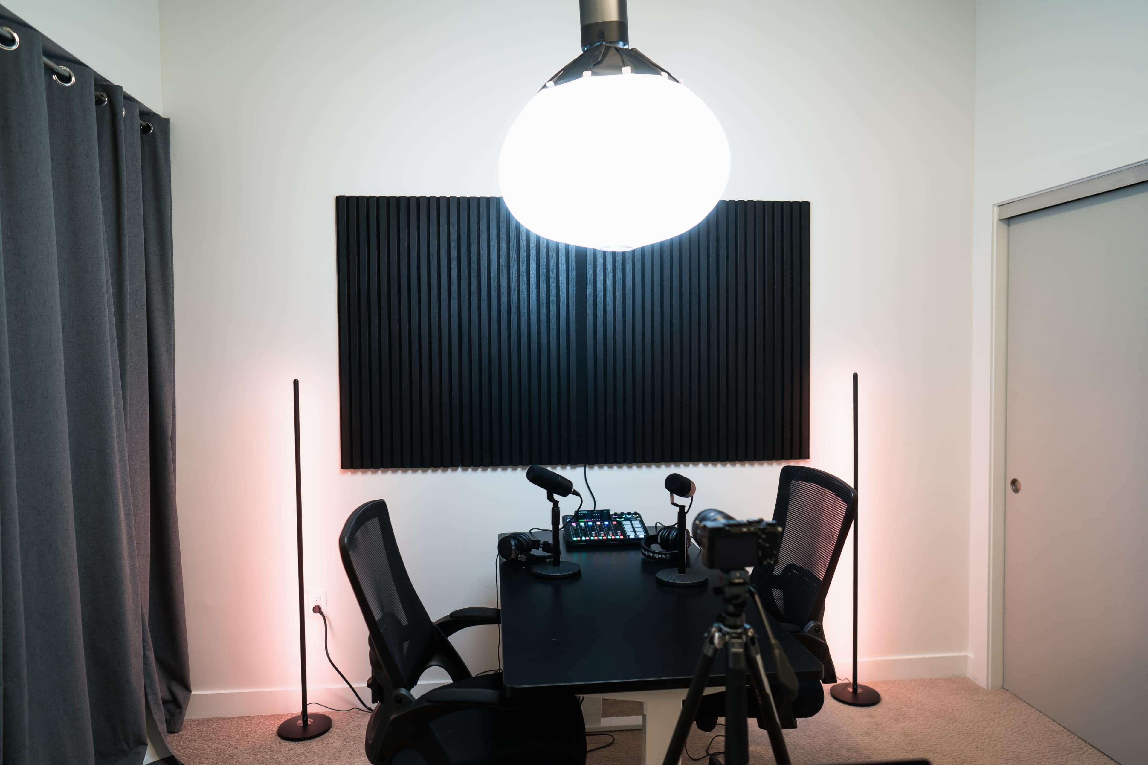 A podcast recording setup features a black table with two chairs, microphones, and a light fixture overhead in a room with soundproofing panels and soft-lit stands on either side.