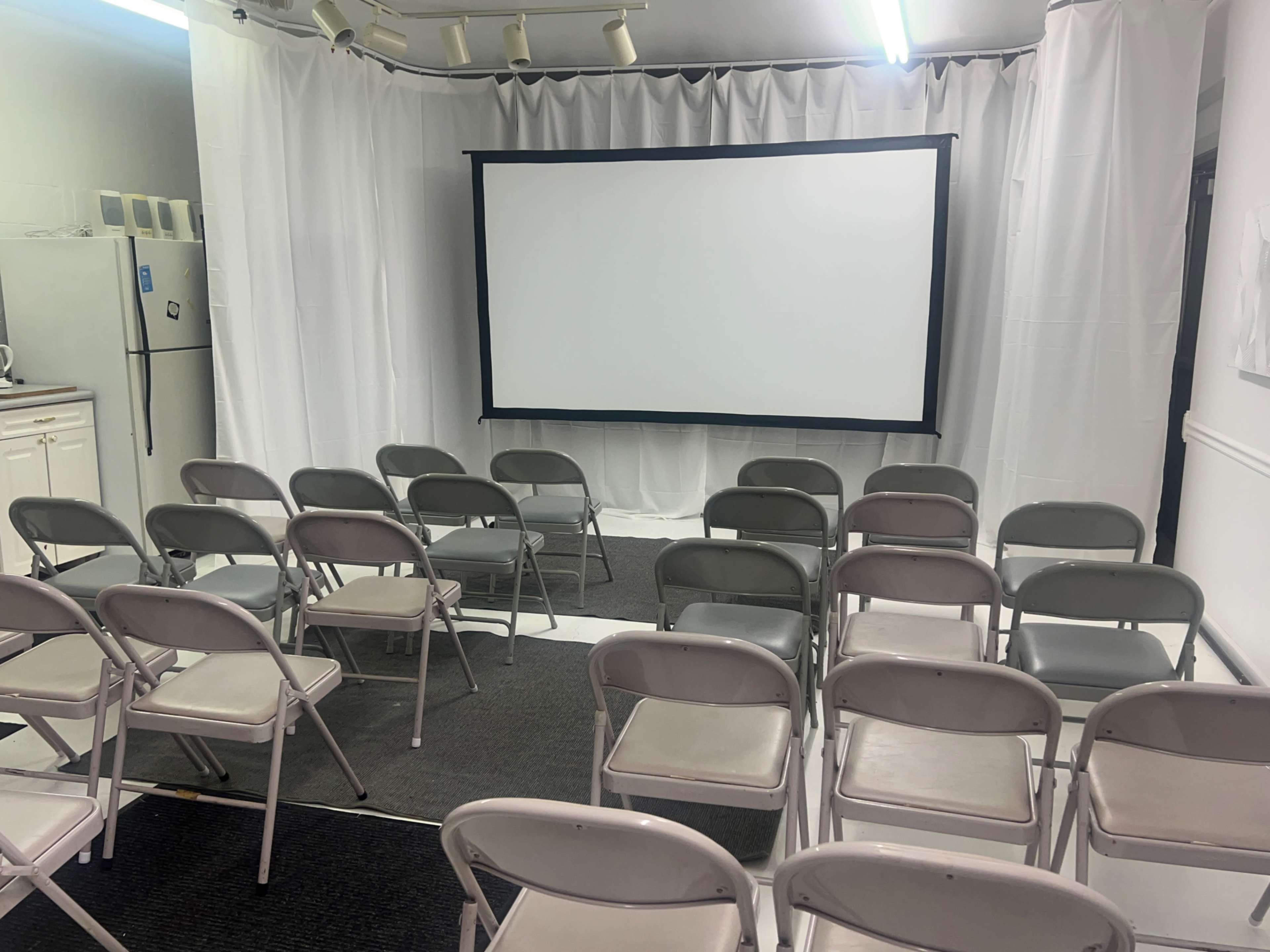 A simple room is set up with a projector screen at the front and several rows of folding chairs arranged in front of it.