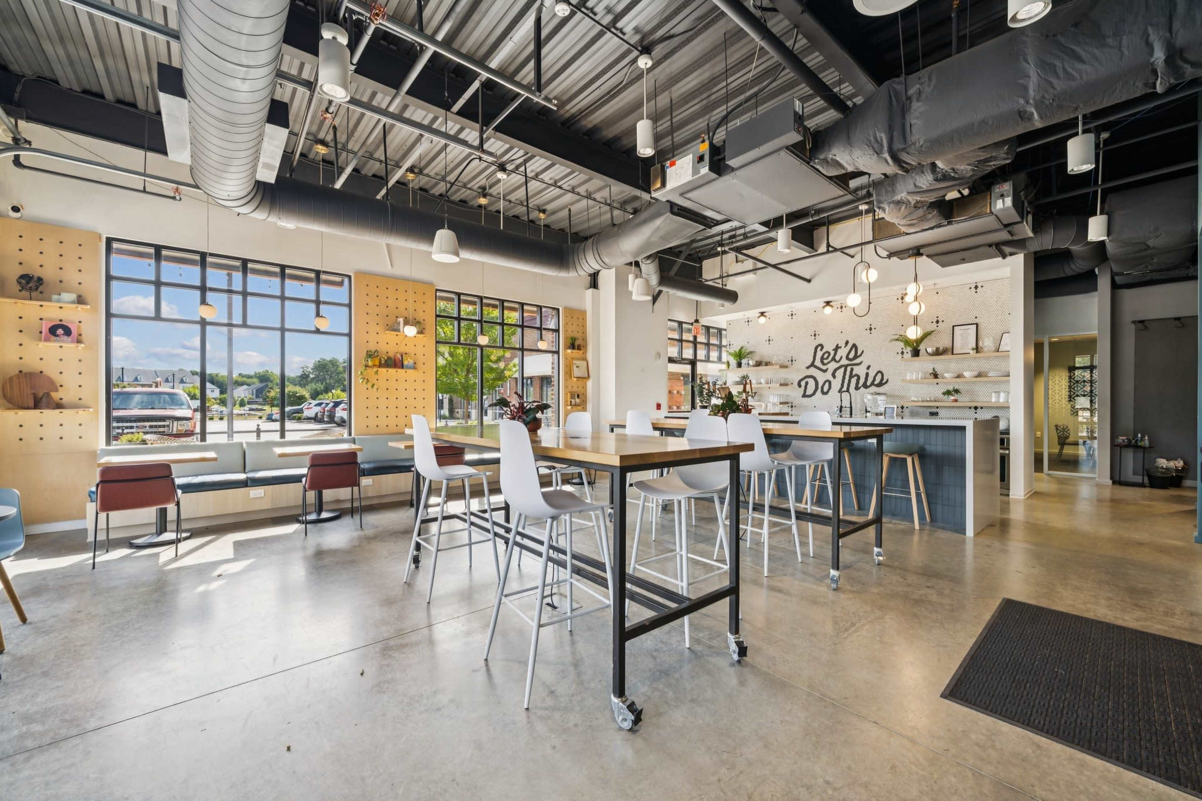 The image shows a modern, open-concept café with tables and chairs arranged around a central area, featuring large windows and a minimalist décor.