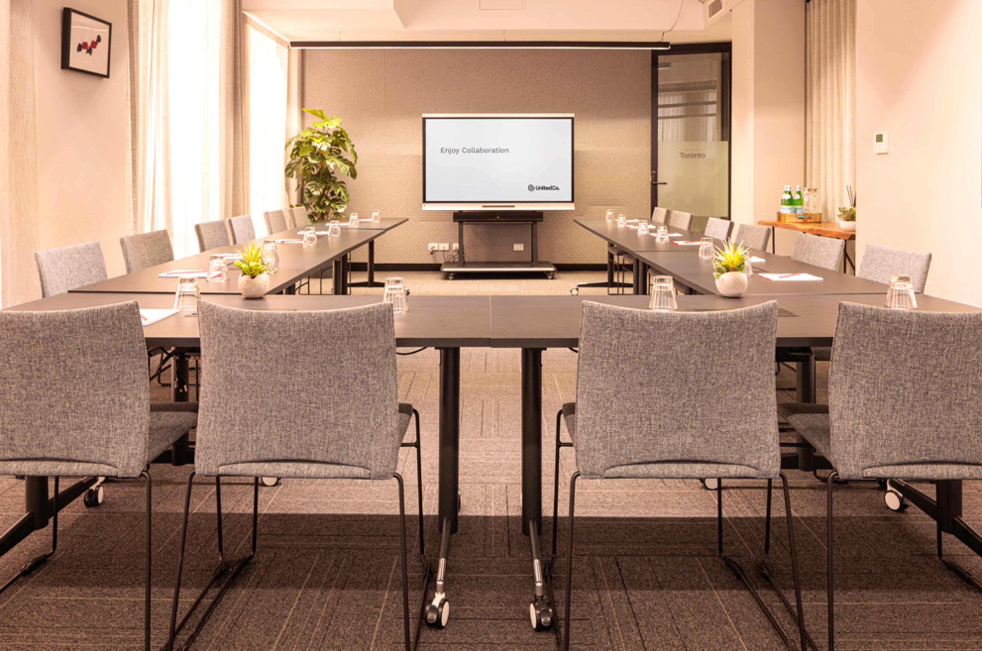 A modern conference room is set up with multiple rectangular tables arranged in a U-shape, each with chairs and water bottles, and features a large screen displaying "Enjoy Collaboration."