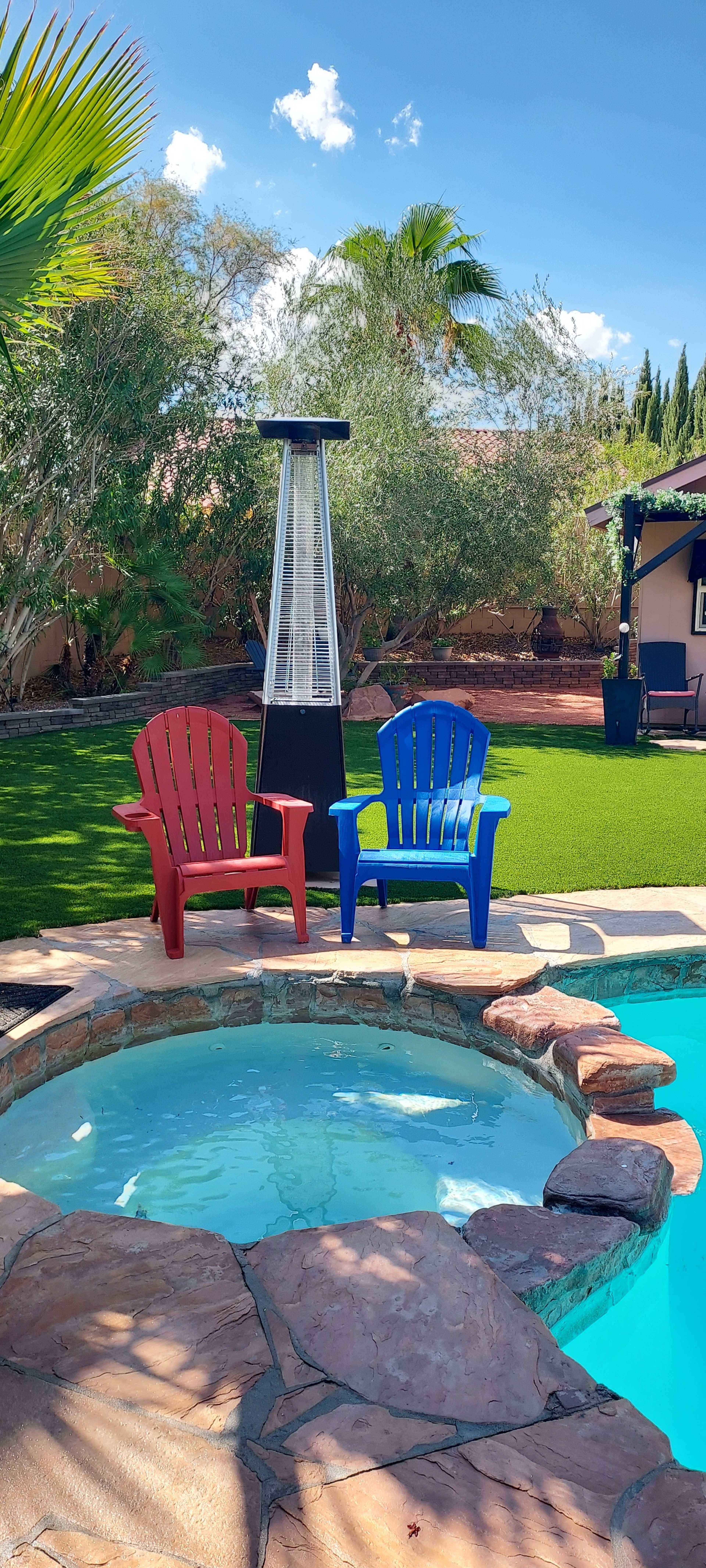 A red chair and a blue chair sit beside a hot tub and a pool in a backyard surrounded by greenery.