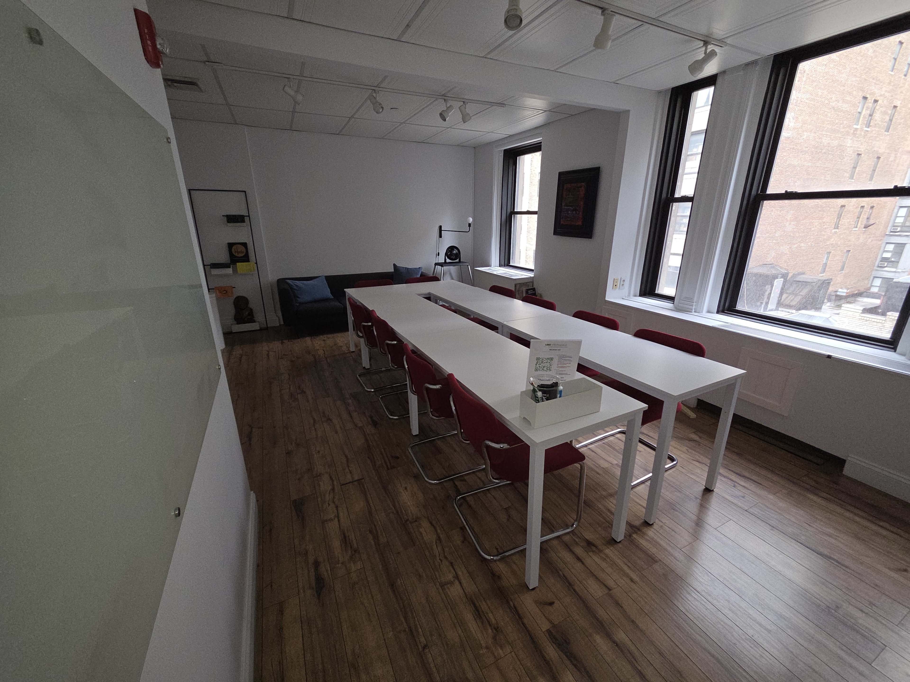 The image shows a spacious meeting room with a long table surrounded by chairs, large windows letting in natural light, and wooden flooring.