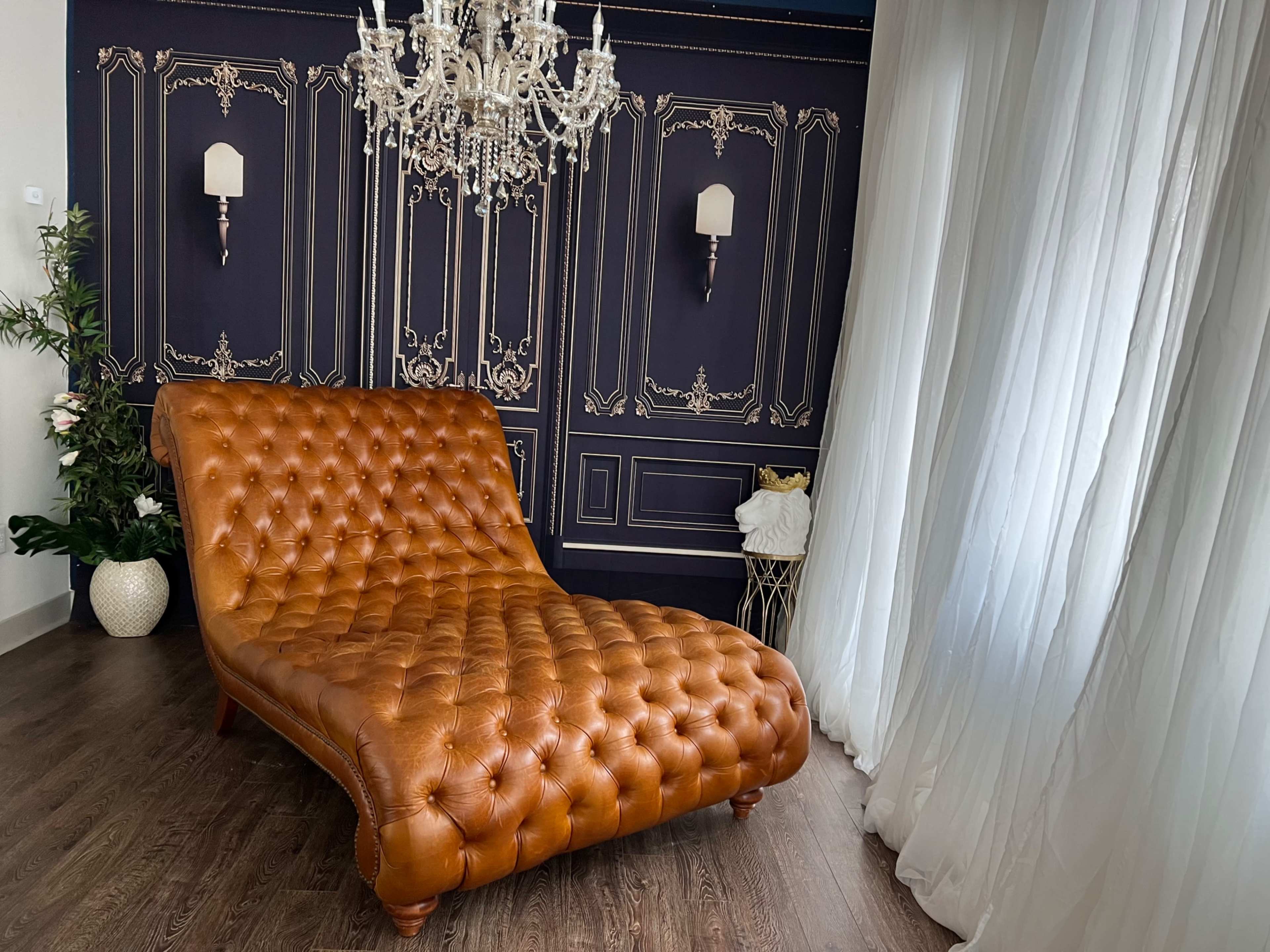 The scene features a tufted brown leather chaise lounge positioned beside sheer white curtains and a decorative wall with a chandelier.