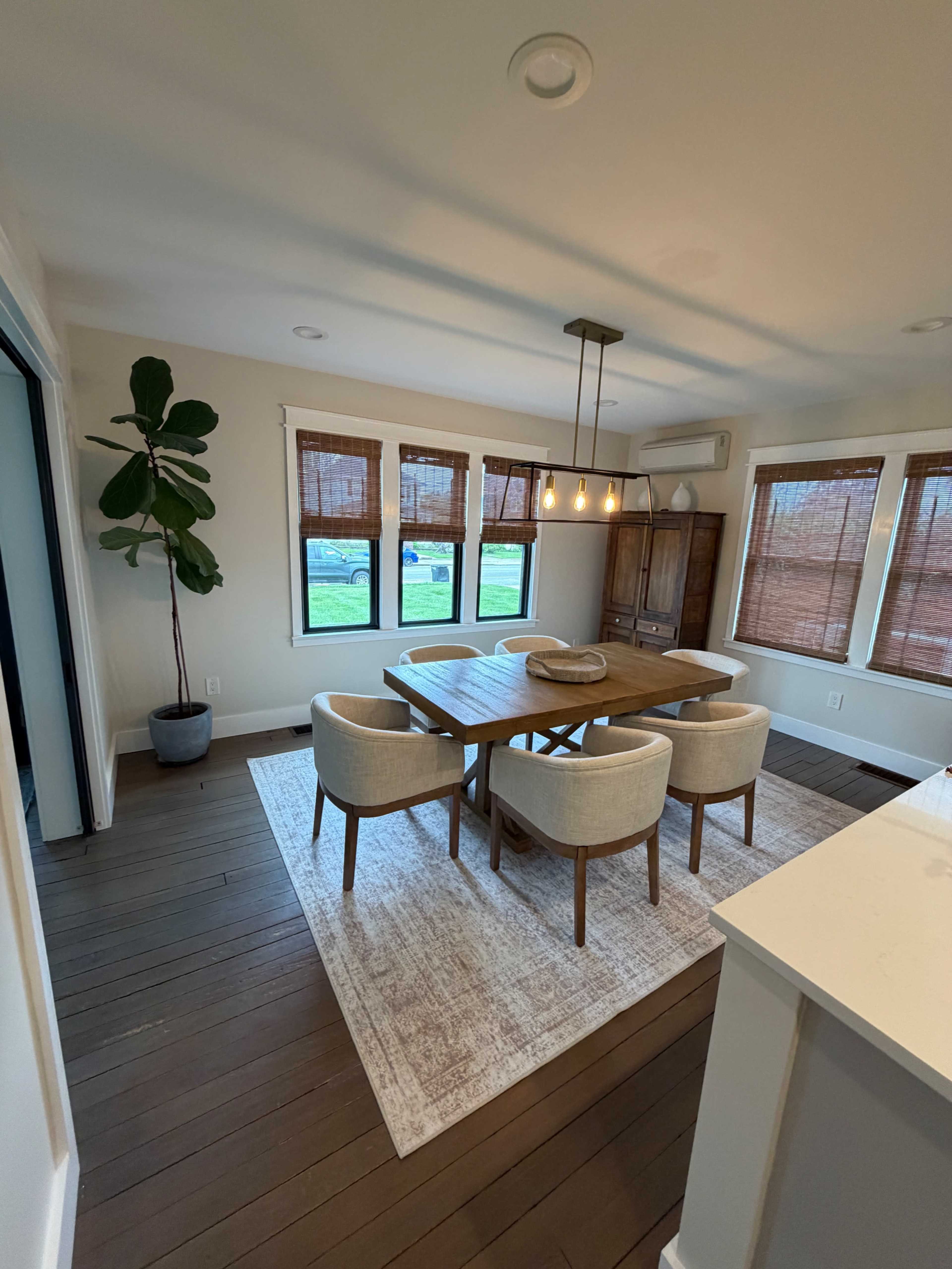 A dining area features a wooden table surrounded by four upholstered chairs, with large windows and a potted plant in the corner.