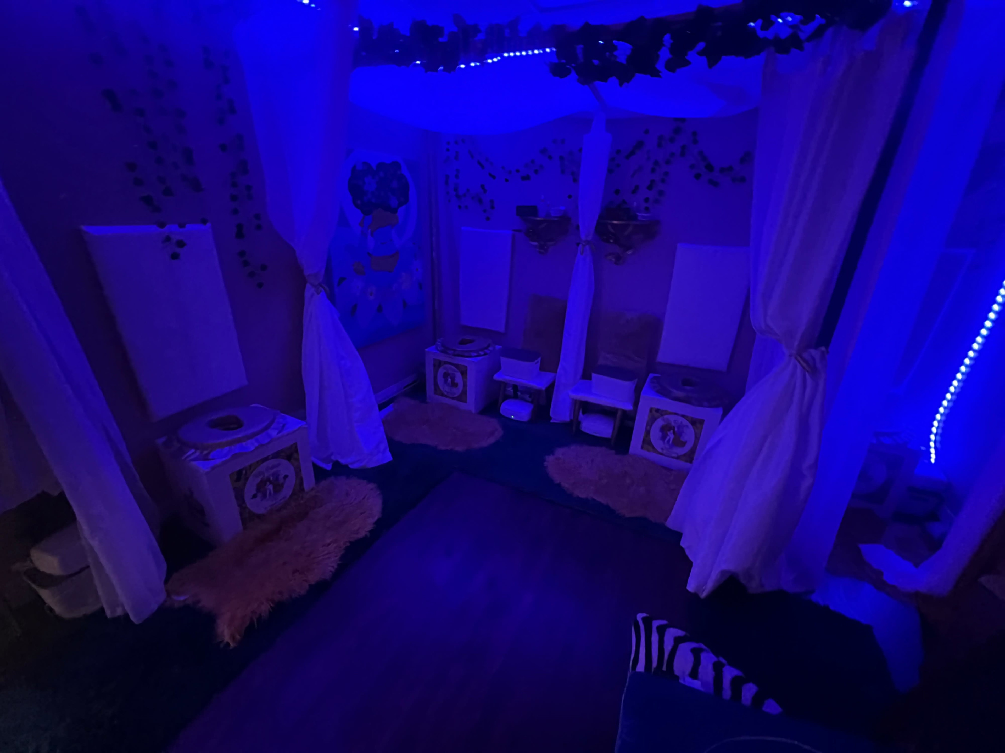 The image shows a dimly lit room with purple lighting, featuring white drapes, furry rugs, and several small seating areas.