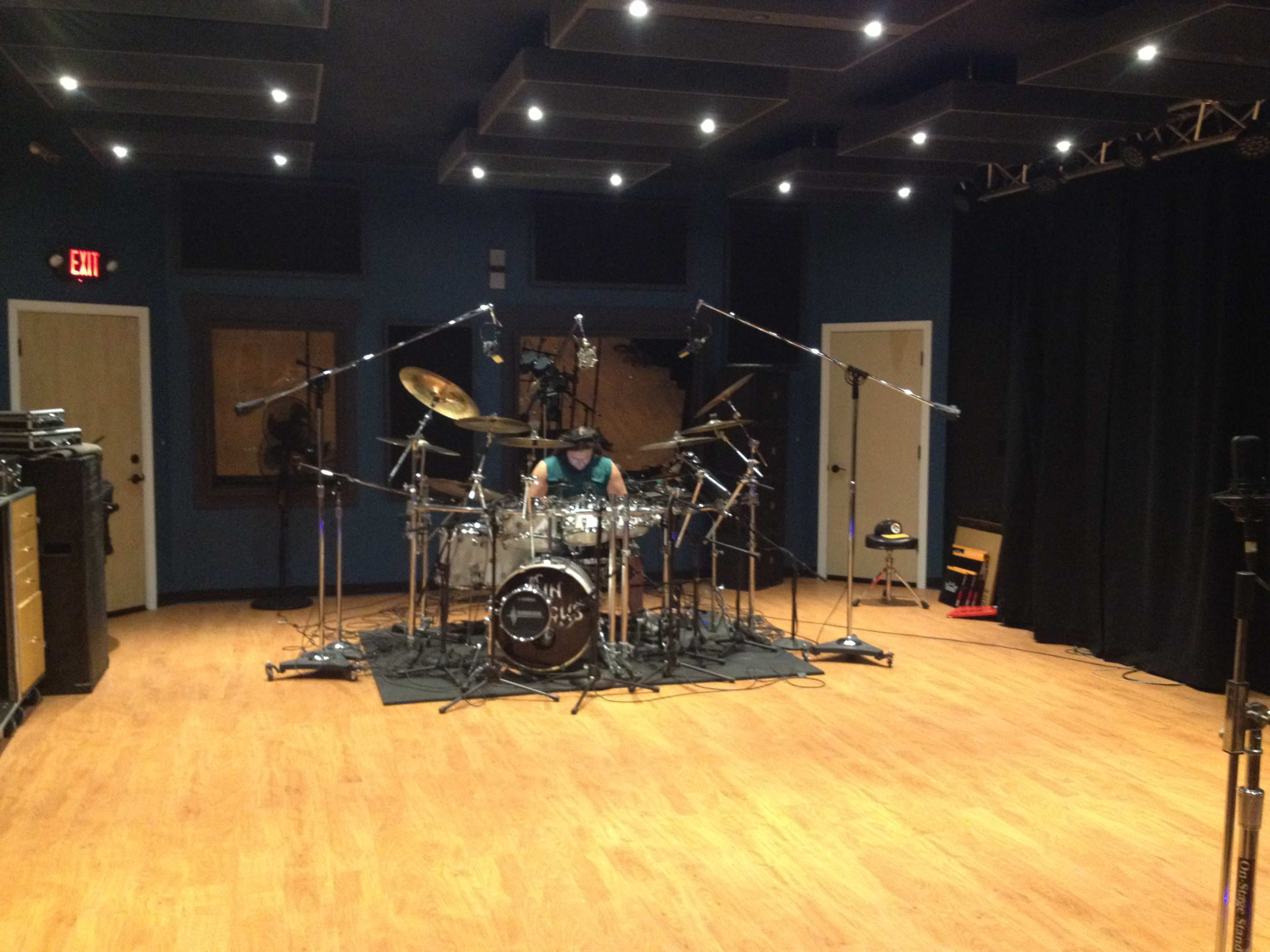 A drummer sits behind a drum set in a spacious recording studio equipped with microphones and soundproofing.
