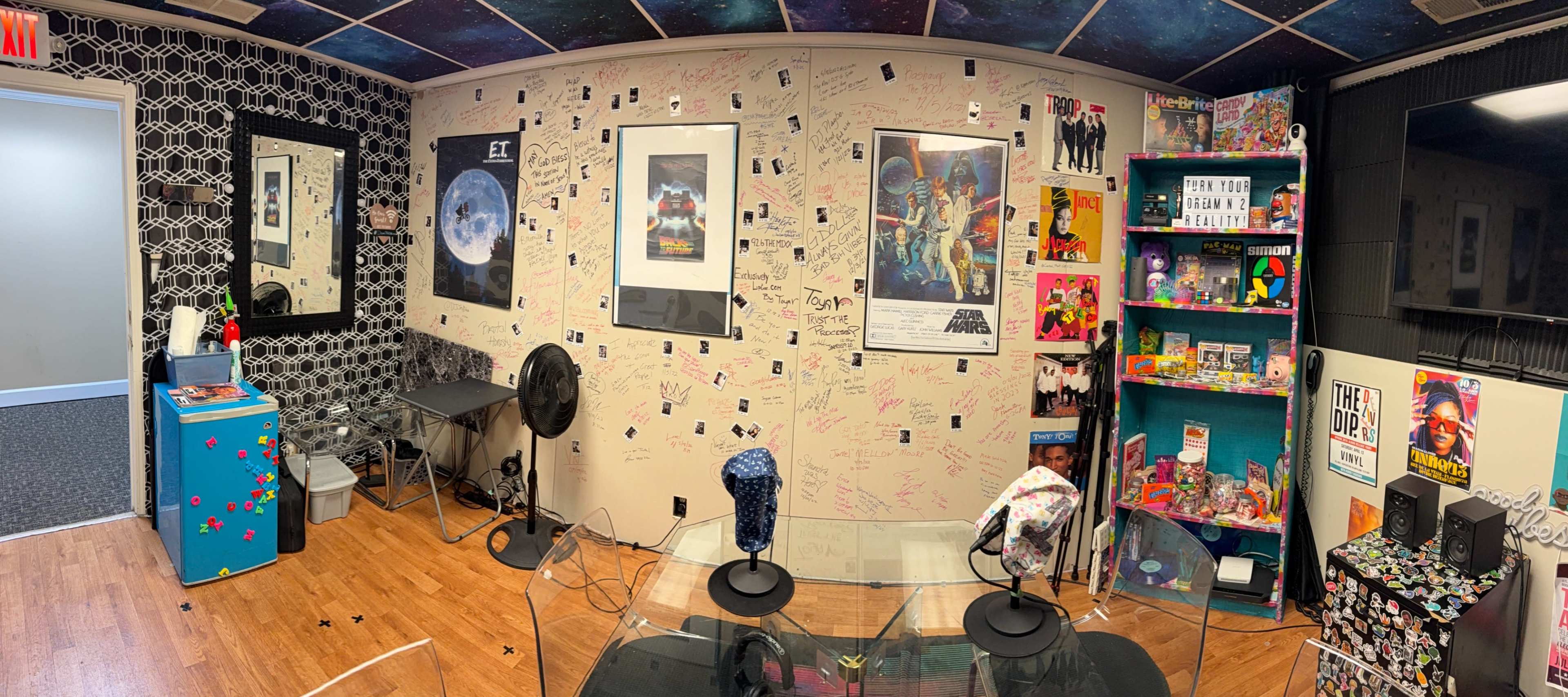 The image shows a colorful room with a glass table, two chairs, walls covered in signatures and drawings, and various posters and shelves filled with toys and memorabilia.