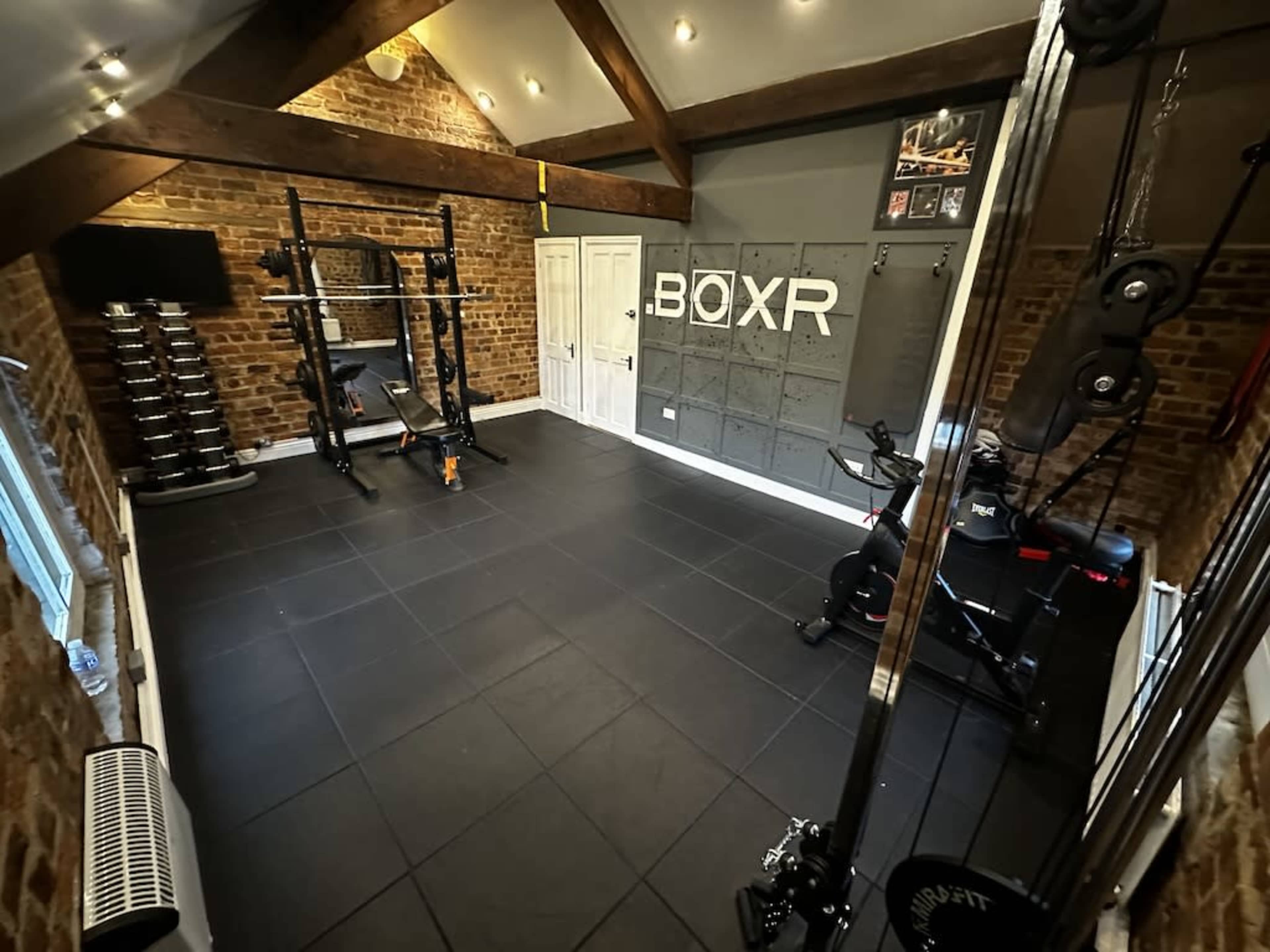 A home gym featuring a weightlifting rack, a stationary bike, various weights, and a cable machine within a room with exposed brick walls and wooden beams.
