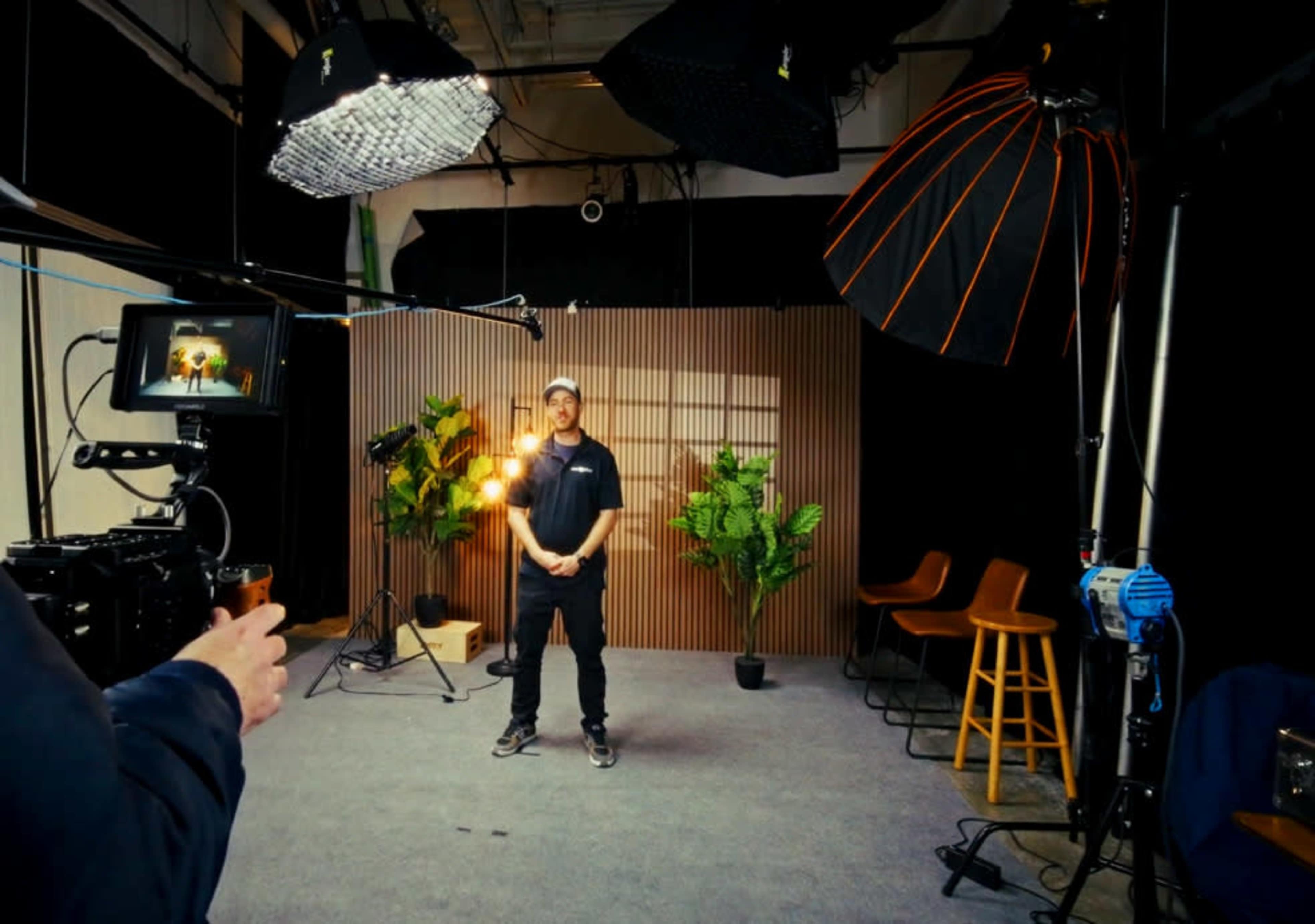 A person stands in a studio setting equipped with professional lighting and cameras, surrounded by potted plants and wooden accents.