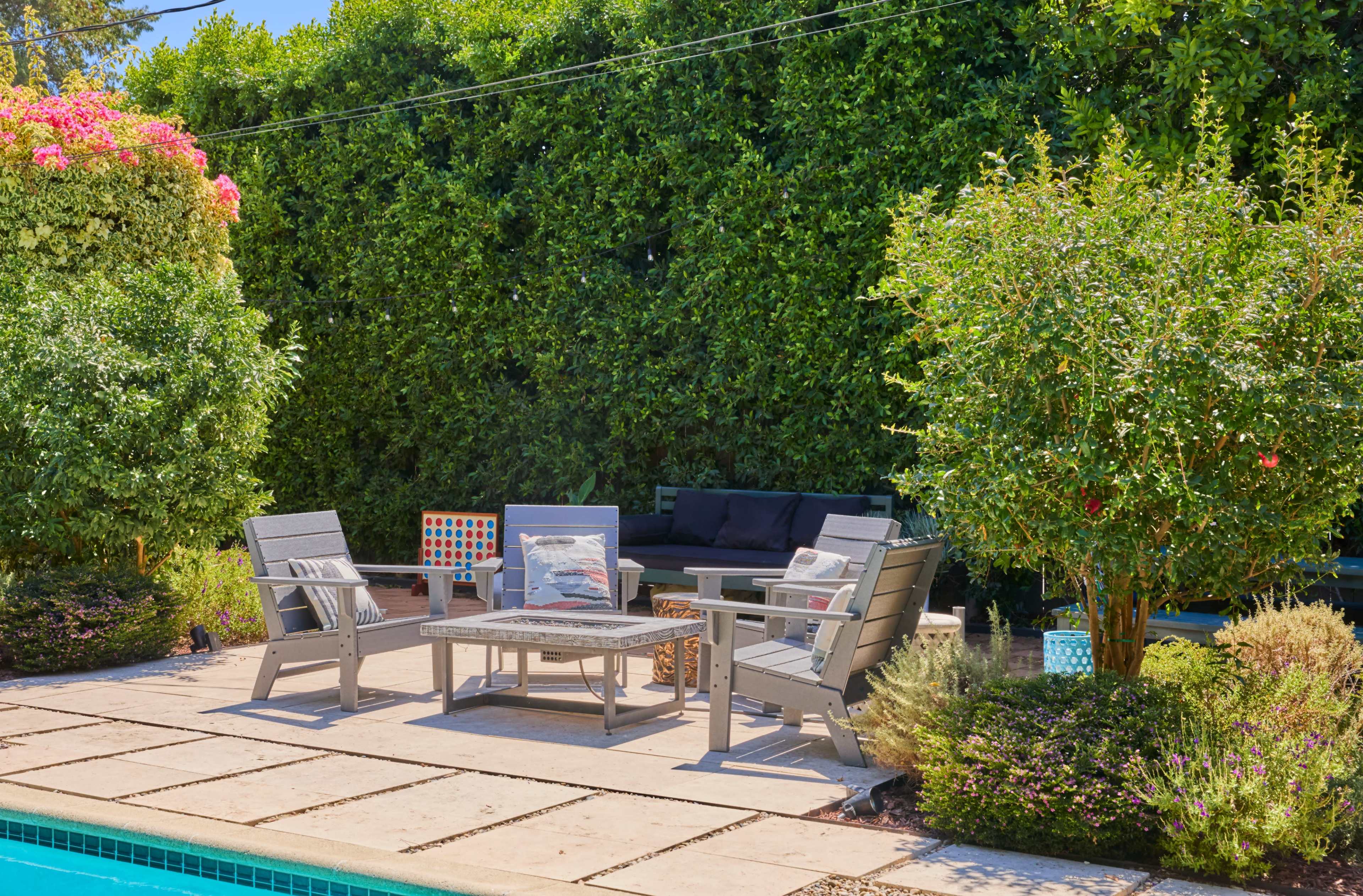 A stone patio with gray outdoor furniture is set near a swimming pool, surrounded by lush greenery and flowering plants.