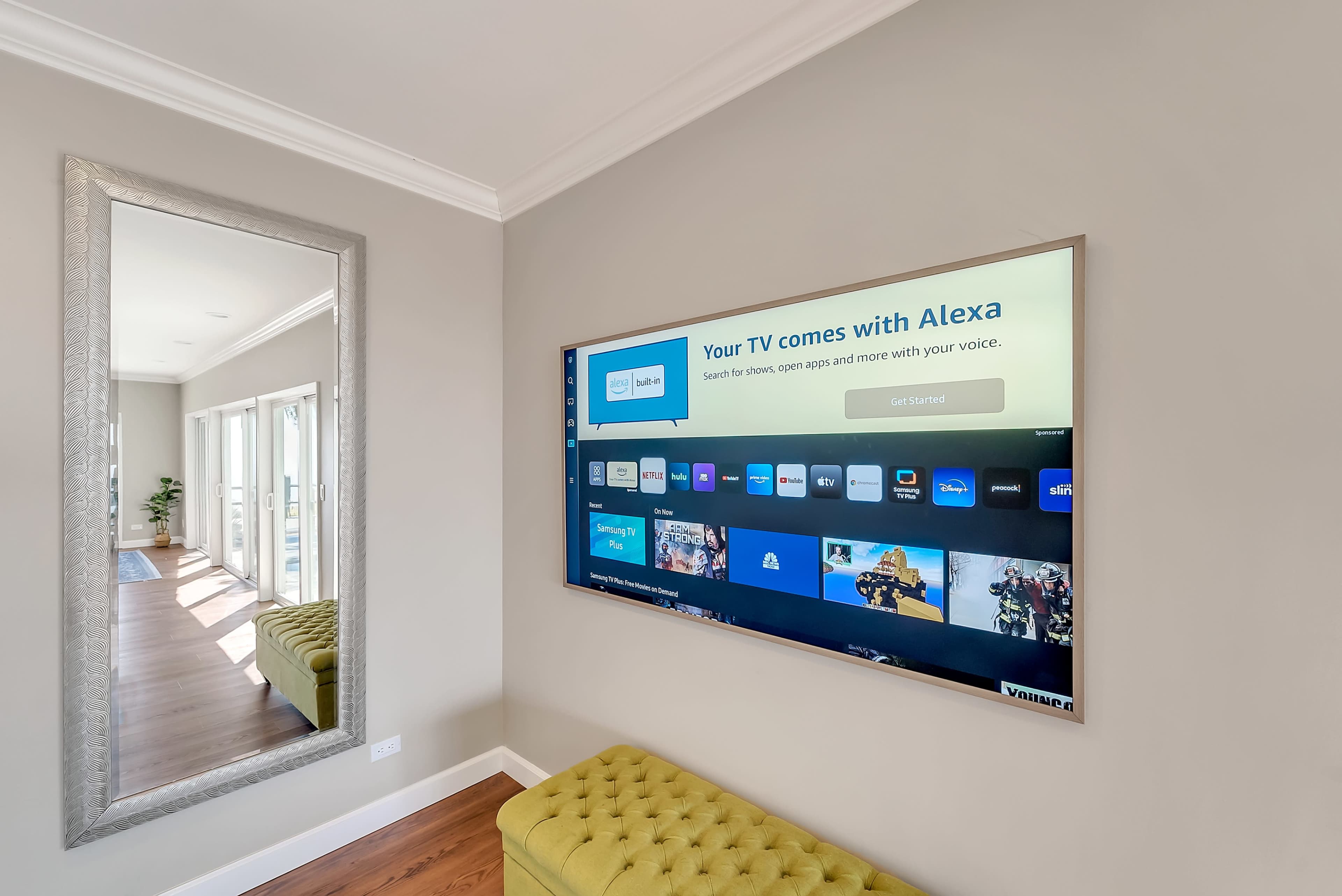 A large flat-screen TV mounted on a wall displays a home screen with various app options, next to a full-length mirror and a yellow tufted bench.