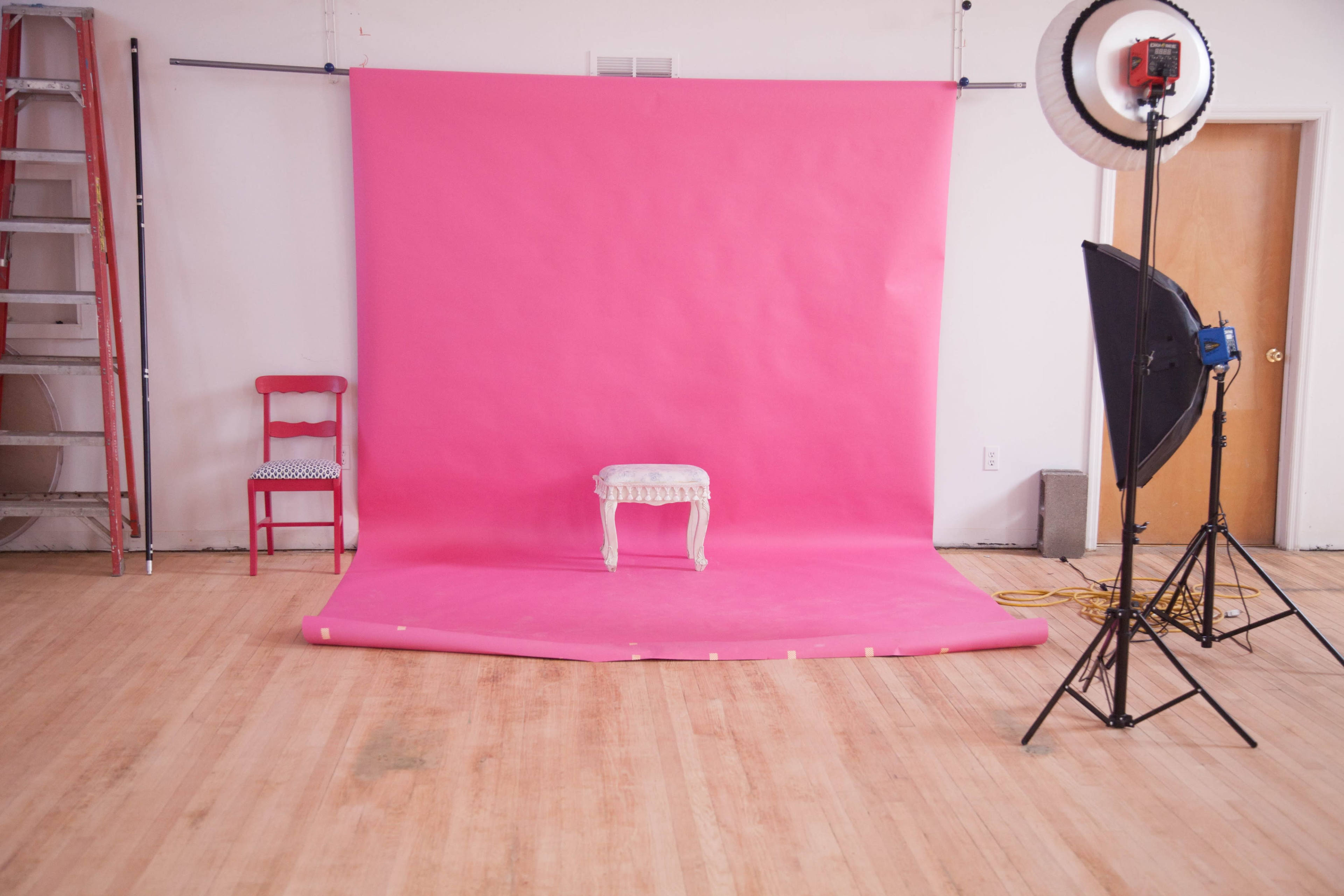 A pink backdrop occupies a photography studio, featuring a small white stool in the center and studio lighting equipment on either side.