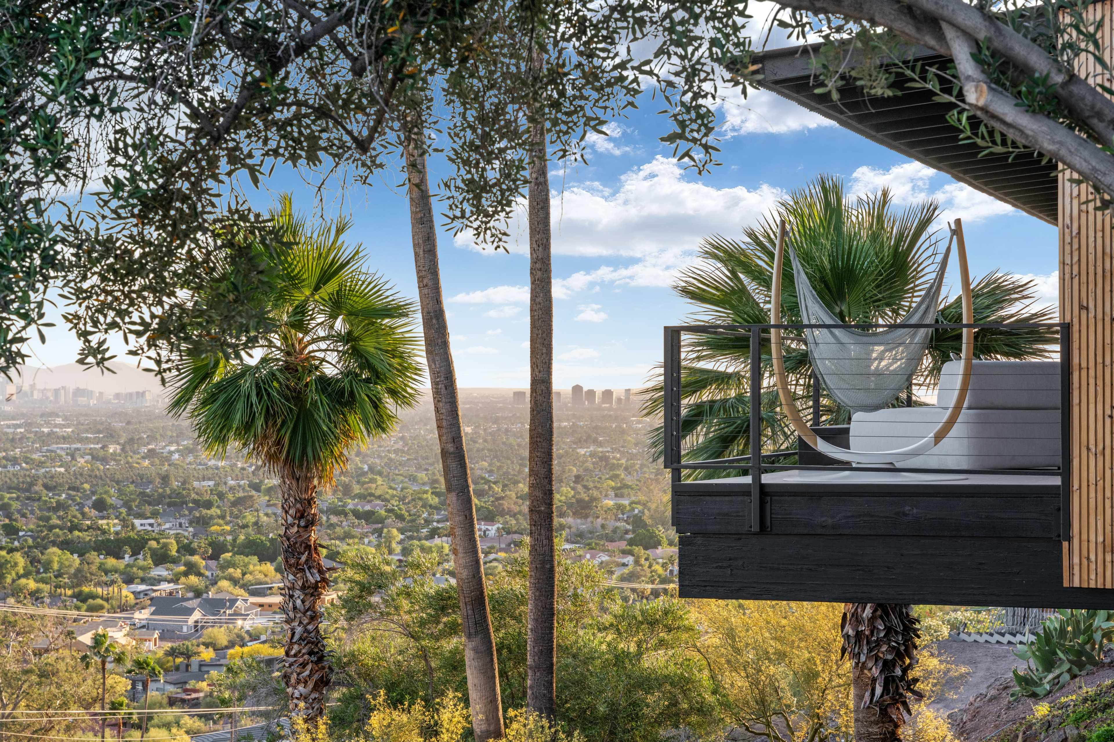 A modern balcony with a hanging chair overlooking a panoramic view of a city skyline surrounded by trees and palm plants.